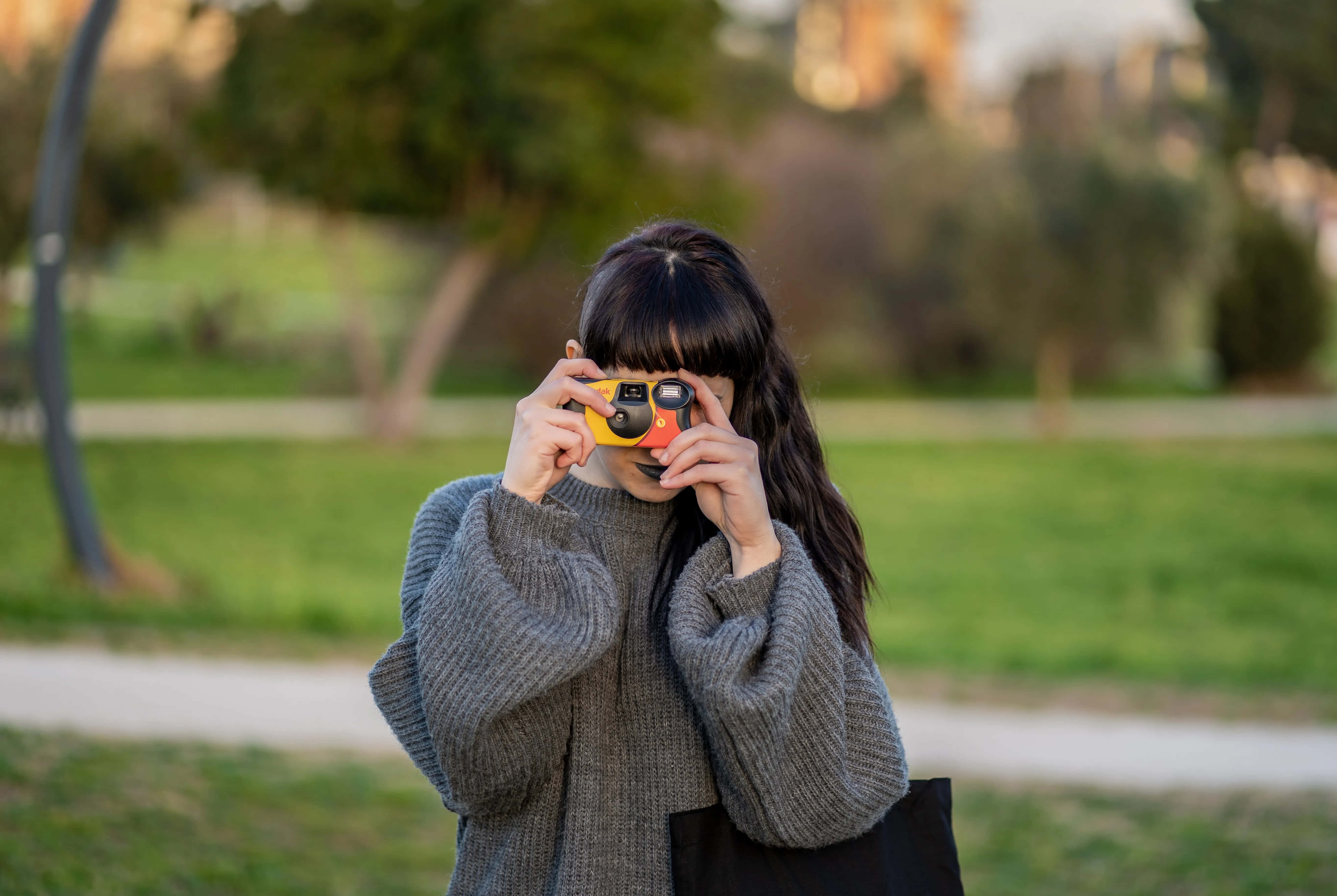 Disposable Camera 桌布 壁紙 — 一位穿著灰色毛衣的年輕女性，在陽光明媚的公園裡，身後是模糊的綠樹和小徑，將一部黃色拍立得相機舉到眼前。