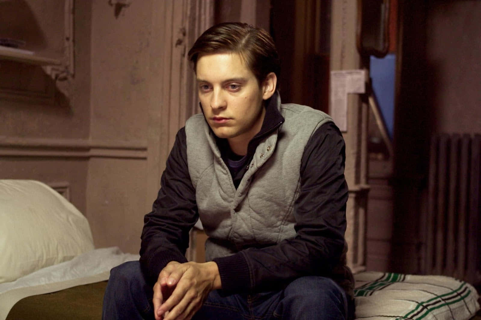 Sad Face 桌布 壁紙 — 一位穿著灰色背心的年輕男子，在光線昏暗的復古房間裡，坐在裝飾牆壁和暖氣片旁的床上沉思。