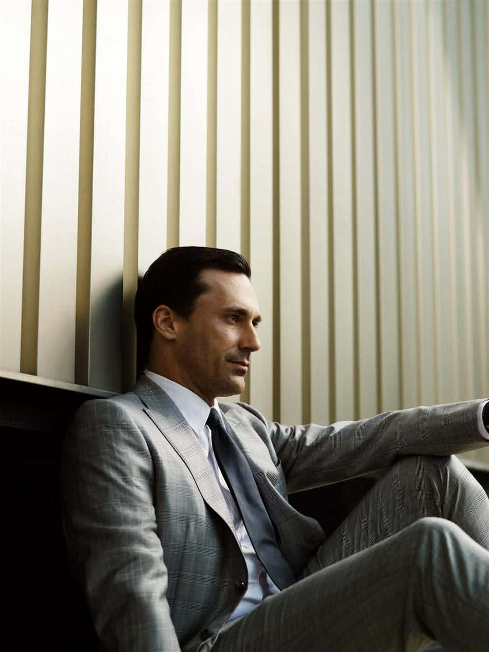 Jon Hamm 桌布 壁紙 — 一位穿著灰色西裝、打扮俐落的男士，在柔和光線的現代室內環境中，靠著垂直百葉窗沉思地坐著。