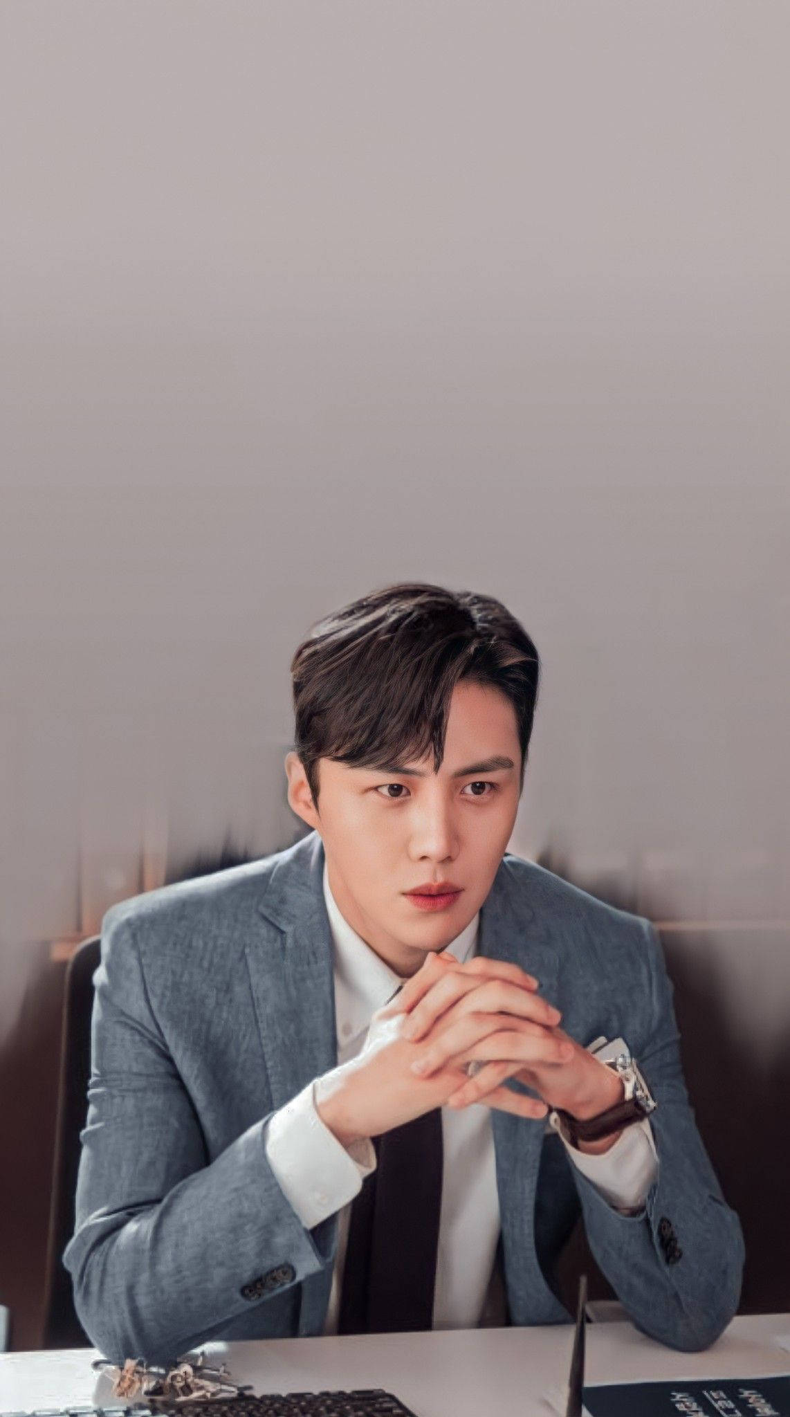 drama 桌布 壁紙 — 一位穿著灰色西裝、衣著講究的男子坐在辦公桌前，雙手緊握，神情嚴肅，背景為柔和的灰色。