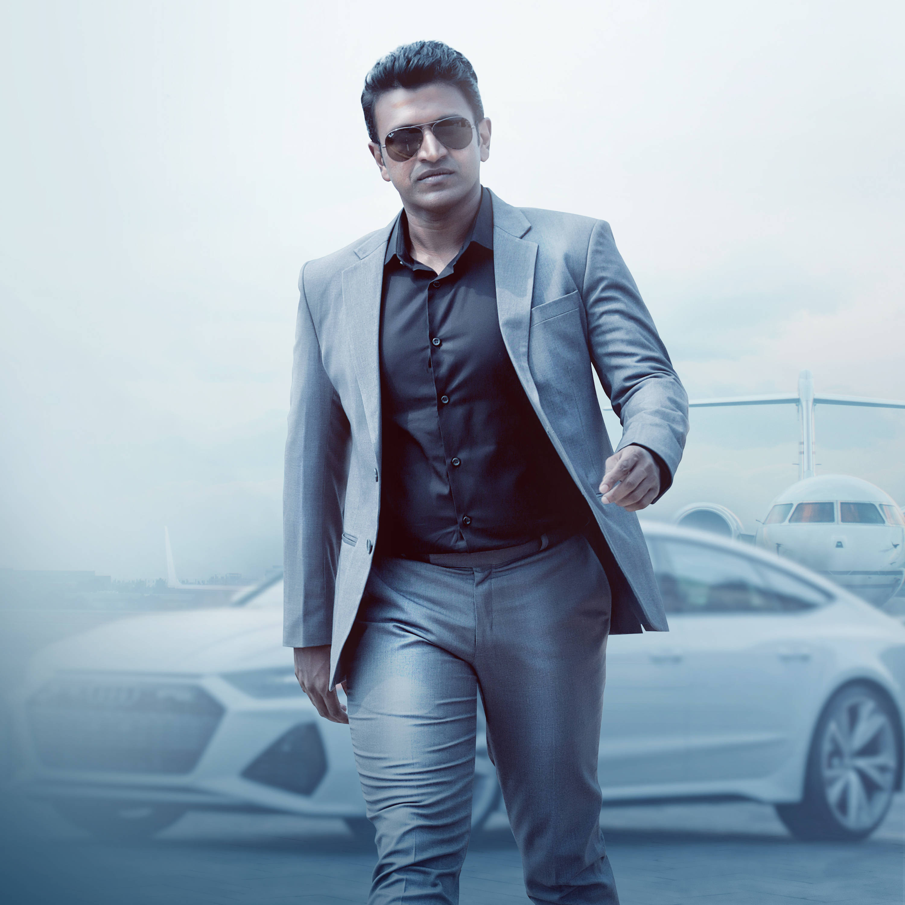 Puneeth Rajkumar 桌布 壁紙 — 一位穿著灰色西裝和太陽眼鏡的時尚男士，自信地走過一輛豪華汽車，背景隱約可見私人飛機。