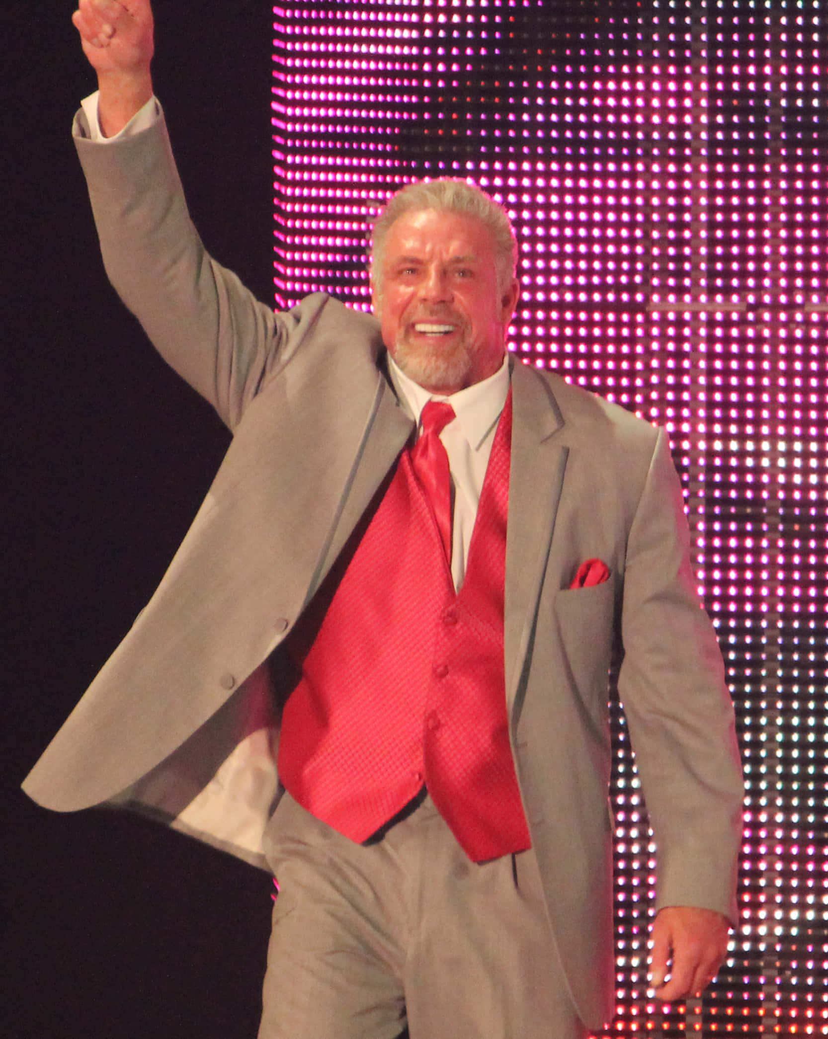 WWE 桌布 壁紙 — 一位穿著灰色西裝和紅色背心的魅力摔角手，在發光的粉紅色LED背景前勝利地舉起拳頭。