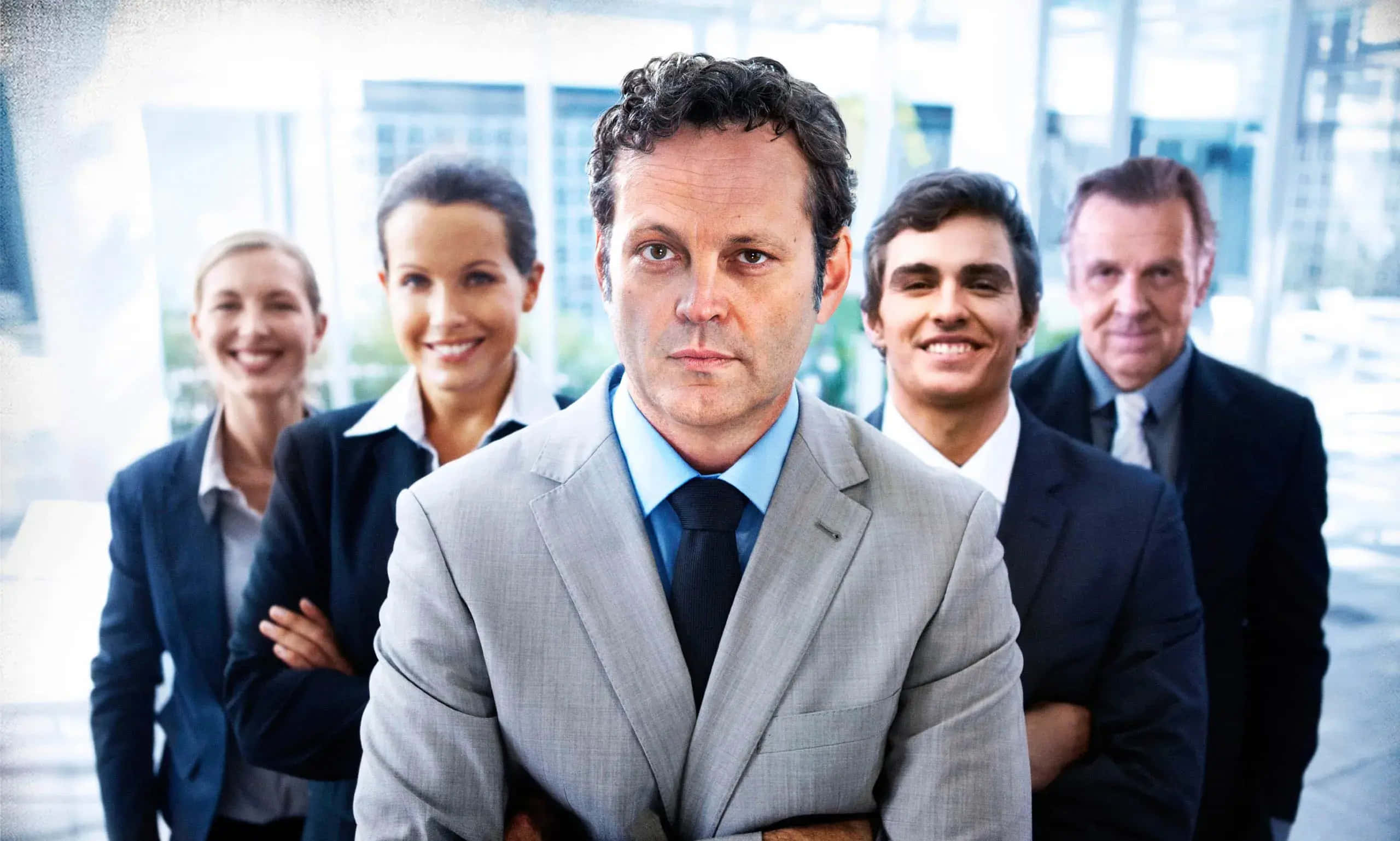 Vince Vaughn 桌布 壁紙 — 一位穿著灰色西裝的嚴肅商人站在前排中央，身後有四位微笑的同事穿著商務服裝，在現代辦公室環境中。