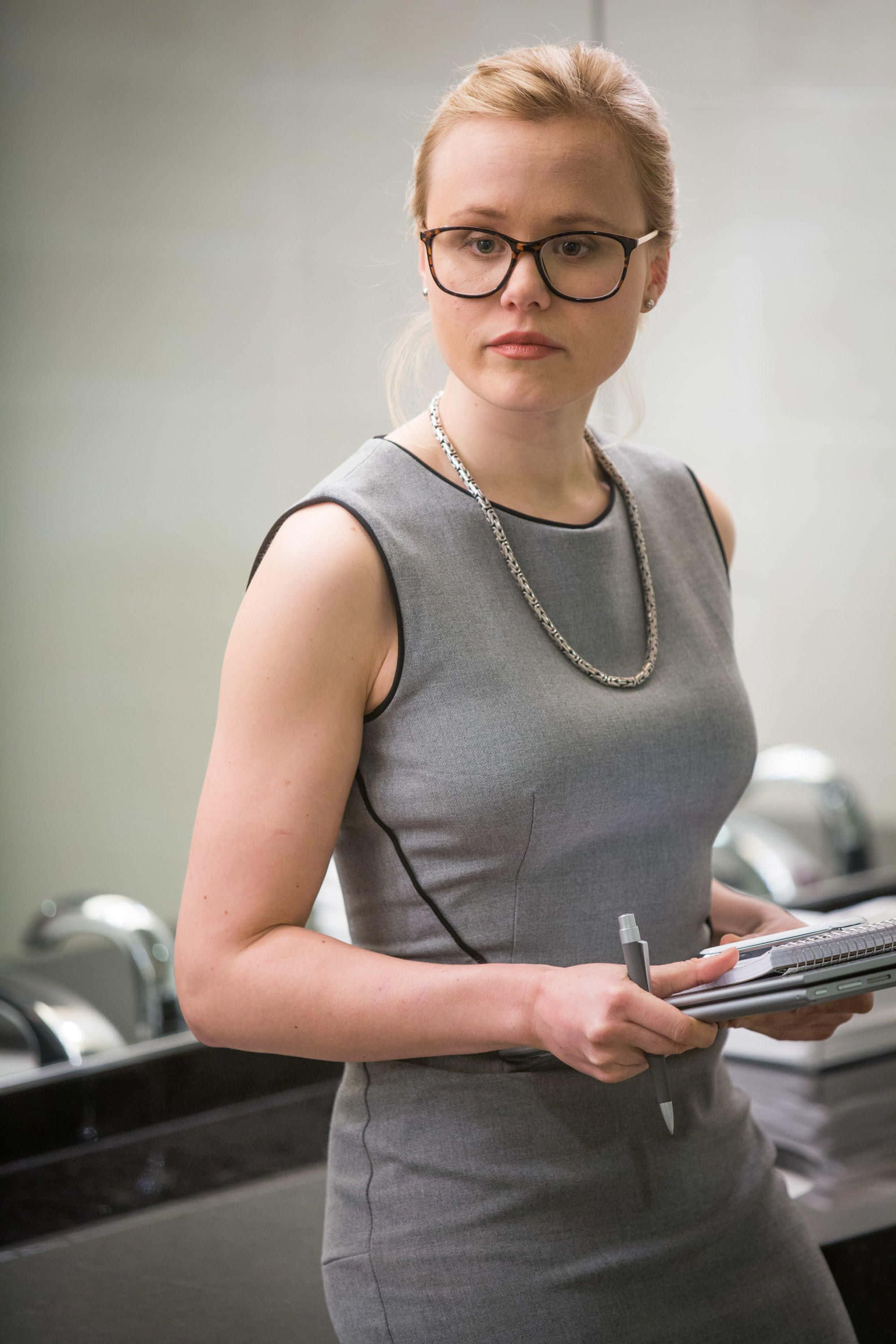 Alison Pill 桌布 壁紙 — 一位穿著灰色連衣裙和眼鏡的女性在現代辦公室環境中拿著筆和記事本。