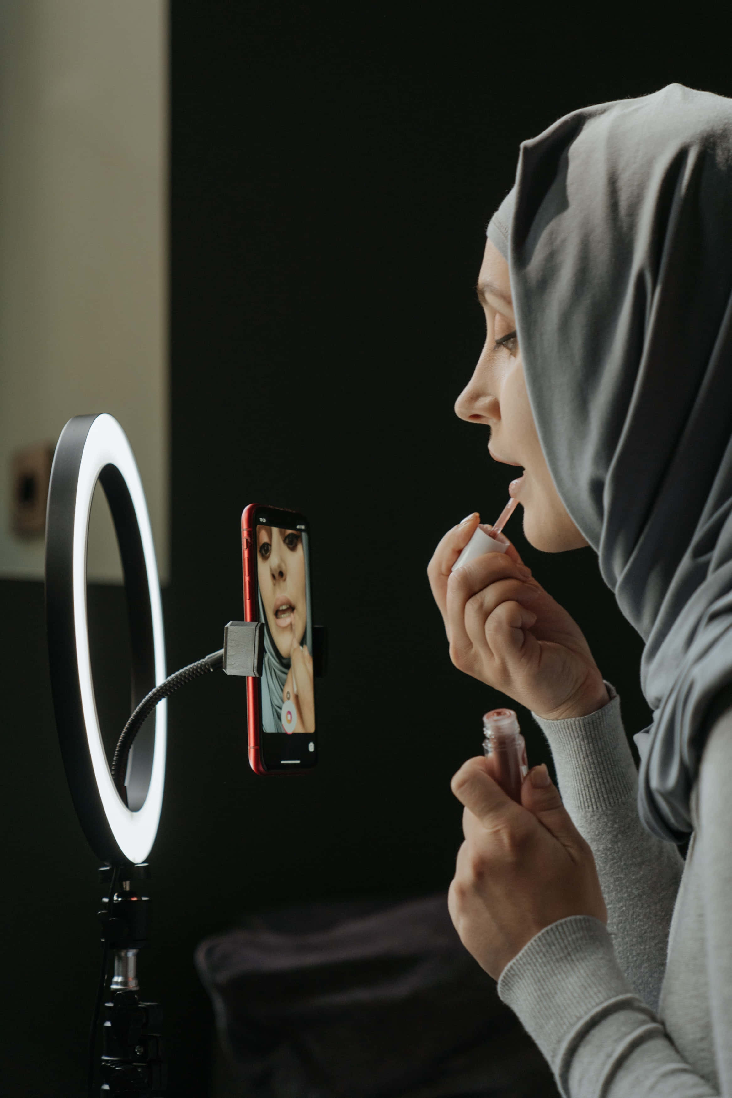Ring Light 桌布 壁紙 — 一位穿著灰色頭巾的女性，一邊看著安裝在明亮環形燈旁邊的智慧型手機上的倒影，一邊塗口紅。
