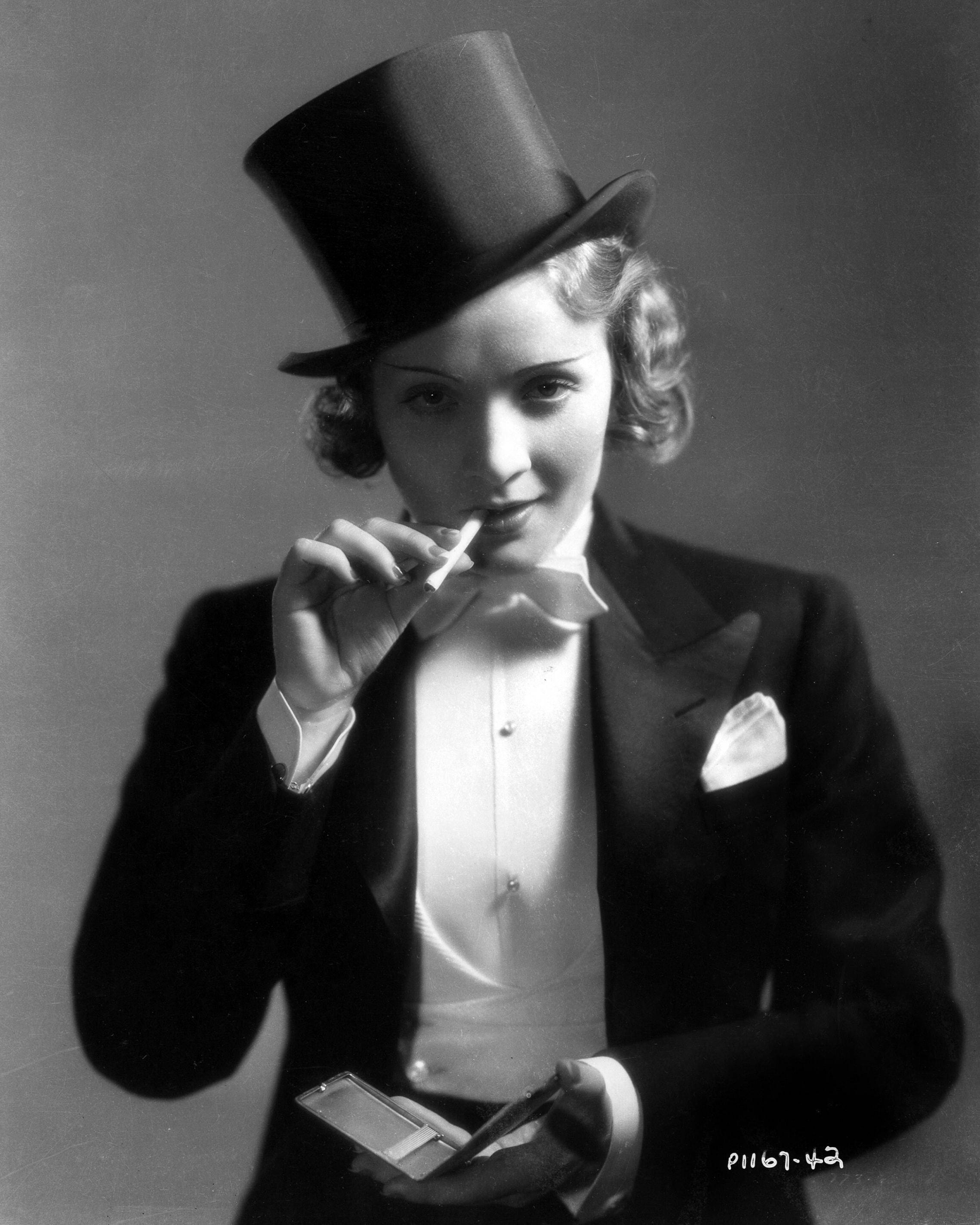 Marlene Dietrich 桌布 壁紙 — 一位穿著燕尾服和高帽的迷人女性，在經典工作室肖像中抽煙，同時拿著一個小鏡子。