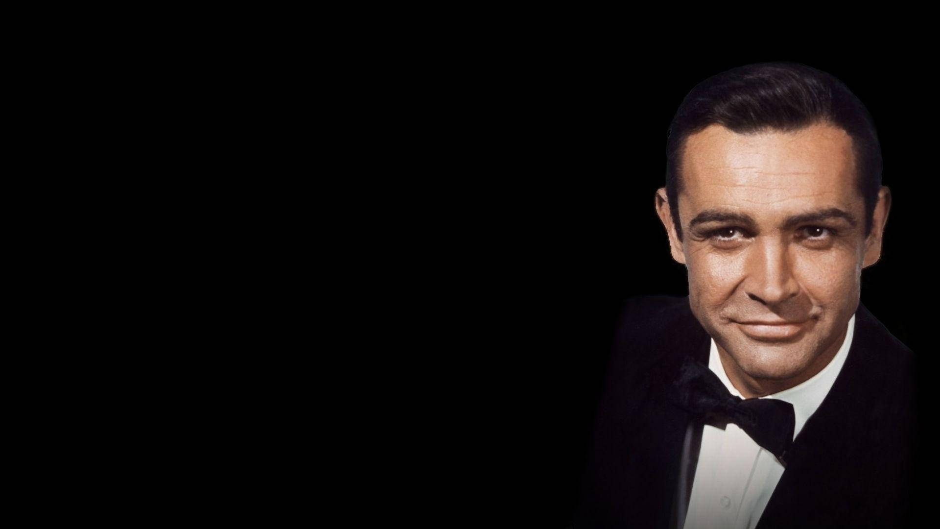 Sean Connery 桌布 壁紙 — 一位穿著燕尾服的男士在純黑背景前露出微妙微笑。