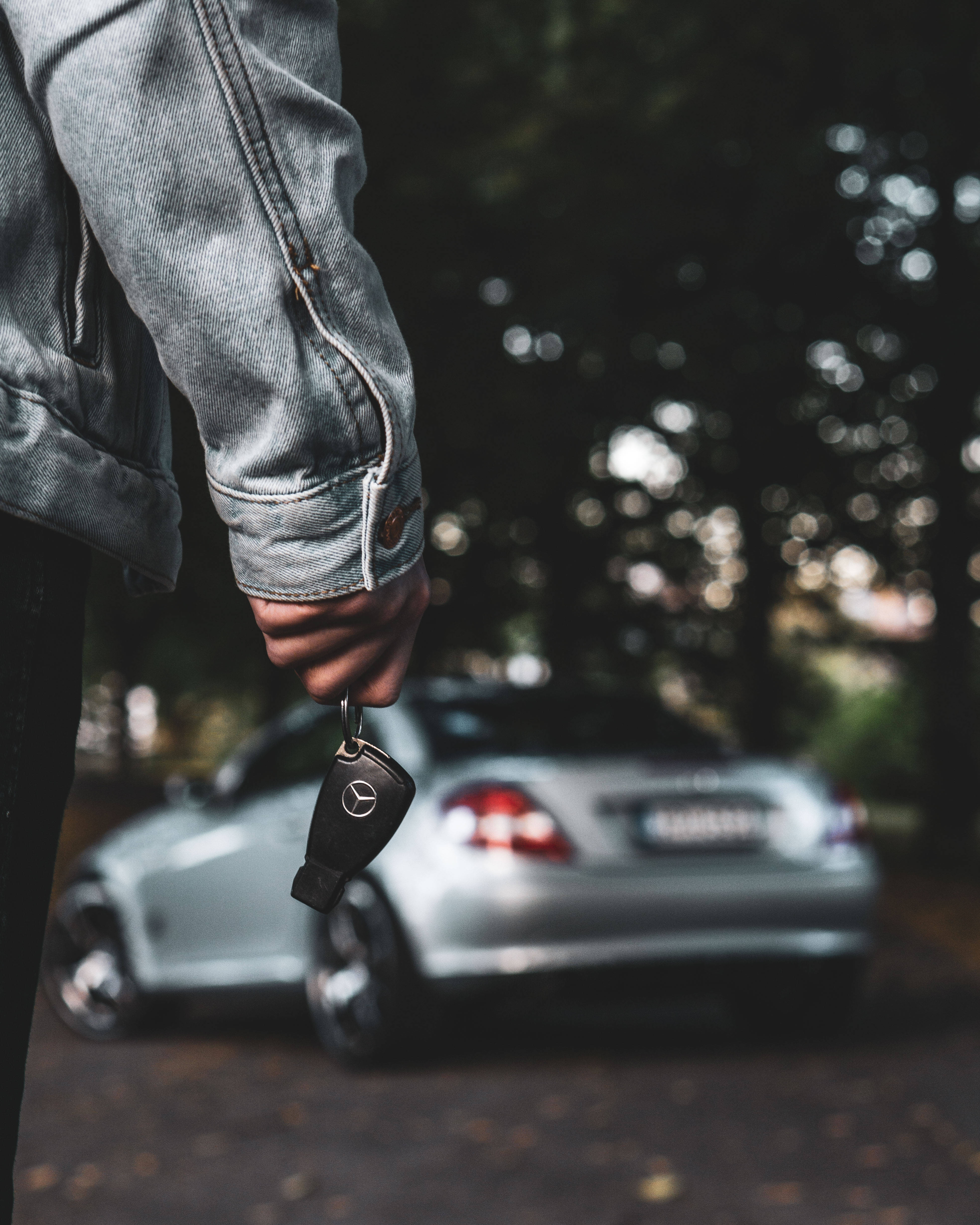 amg iPhone 桌布 壁紙 — 一位穿著牛仔外套的人手持賓士車鑰匙，背景有一輛銀色汽車在柔和晚燈下模糊不清。