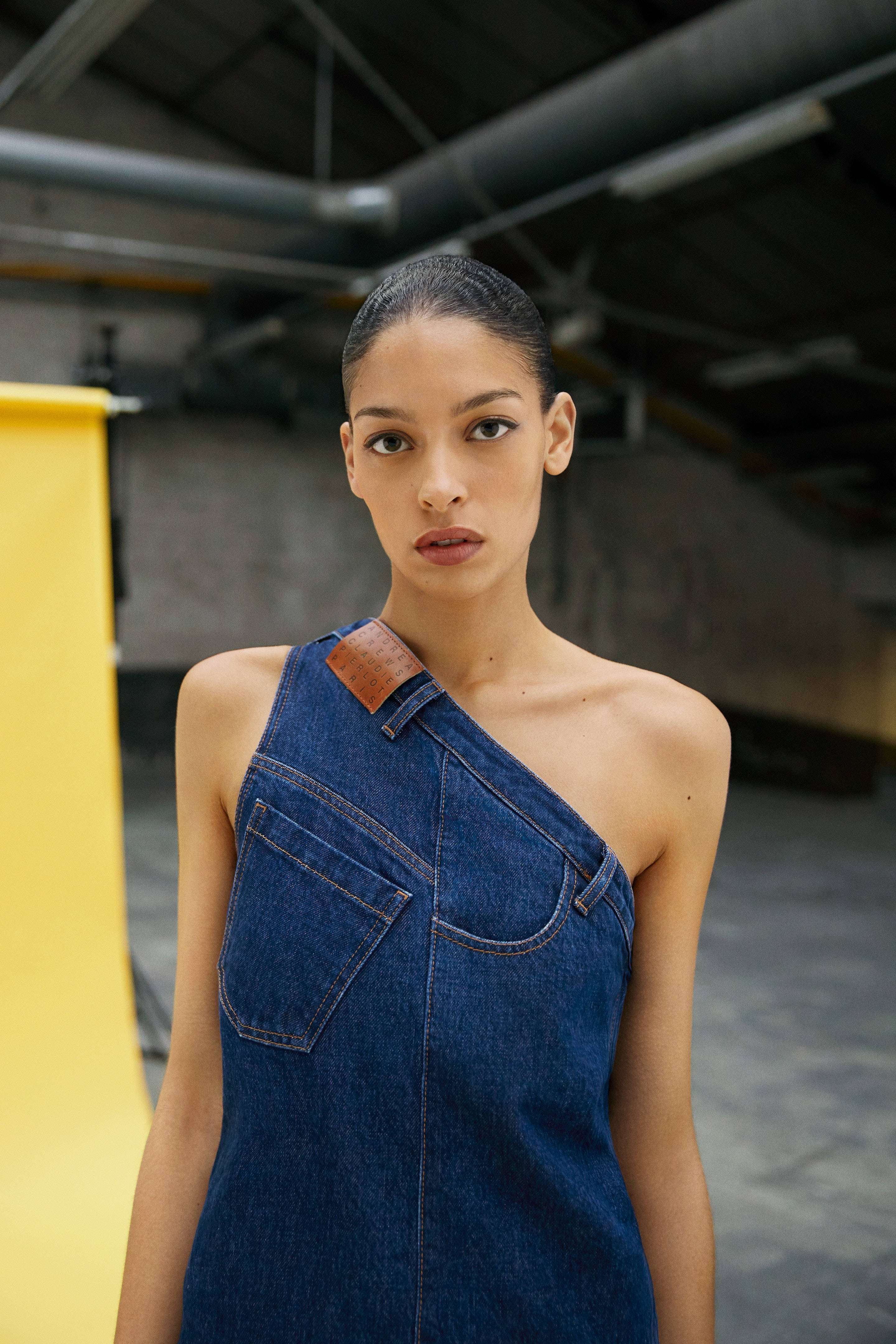 Denim 桌布 壁紙 — 一位穿著牛仔布單肩連衣裙的優雅女士，自信地站在一個有裸露管道和鮮豔黃色背景的工業倉庫中。