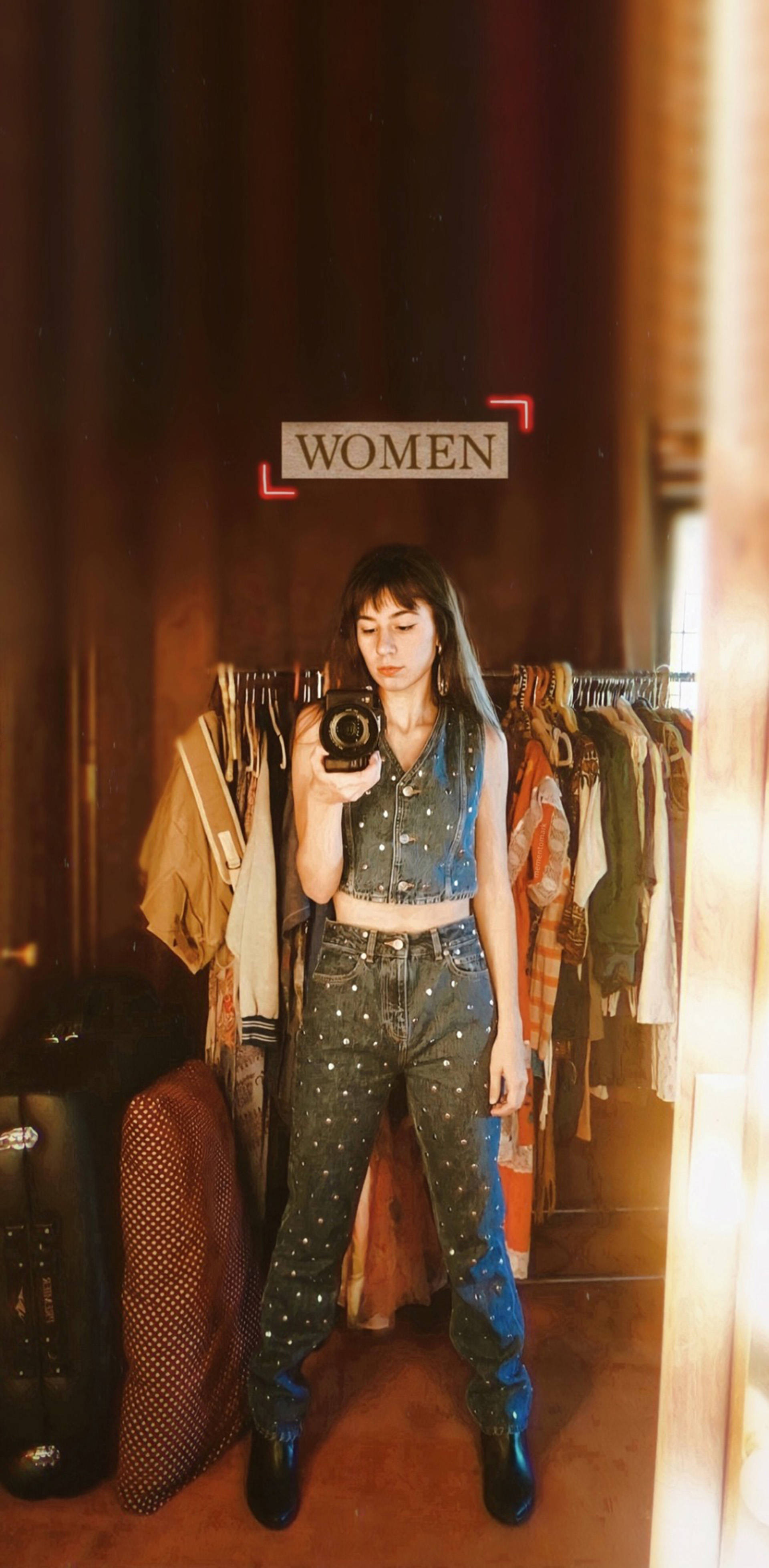 桌布 壁紙 — 一位穿著牛仔布服裝的女性在燈光溫暖的服裝店內，在貨架上方有「WOMEN」標誌的情況下，對著鏡子自拍。