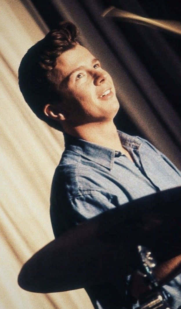 Rick Astley 桌布 壁紙 — 一位穿著牛仔襯衫的年輕男子，在垂簾背景前打鼓時向上微笑。