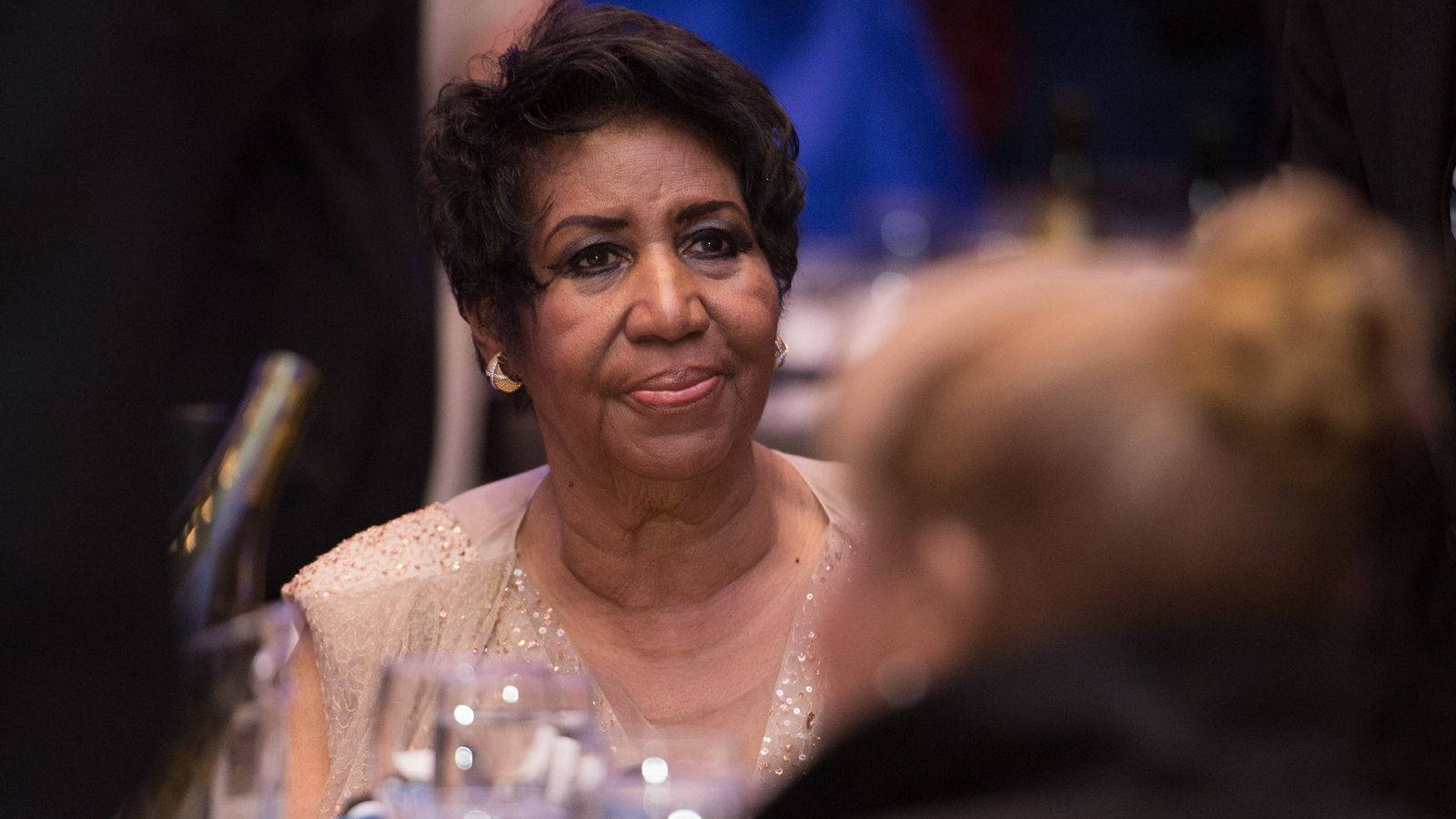 Celebrity 桌布 壁紙 — 一位穿著珠飾長裙的優雅女性，坐在正式的晚餐桌旁，周圍有酒杯和模糊的賓客，她溫暖地微笑。
