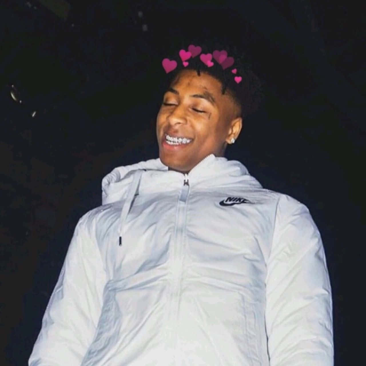  壁紙 — 一位穿著白色Nike外套的年輕男子在黑暗背景上，頭頂飄浮著粉紅色心形表情符號，開心地微笑。