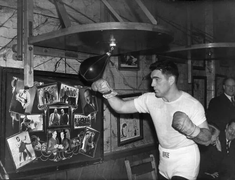 Boxing 桌布 壁紙 — 一位穿著白色短褲和手套的拳擊手在一個光線昏暗、掛滿裱框照片的健身房中擊打重型沙包。