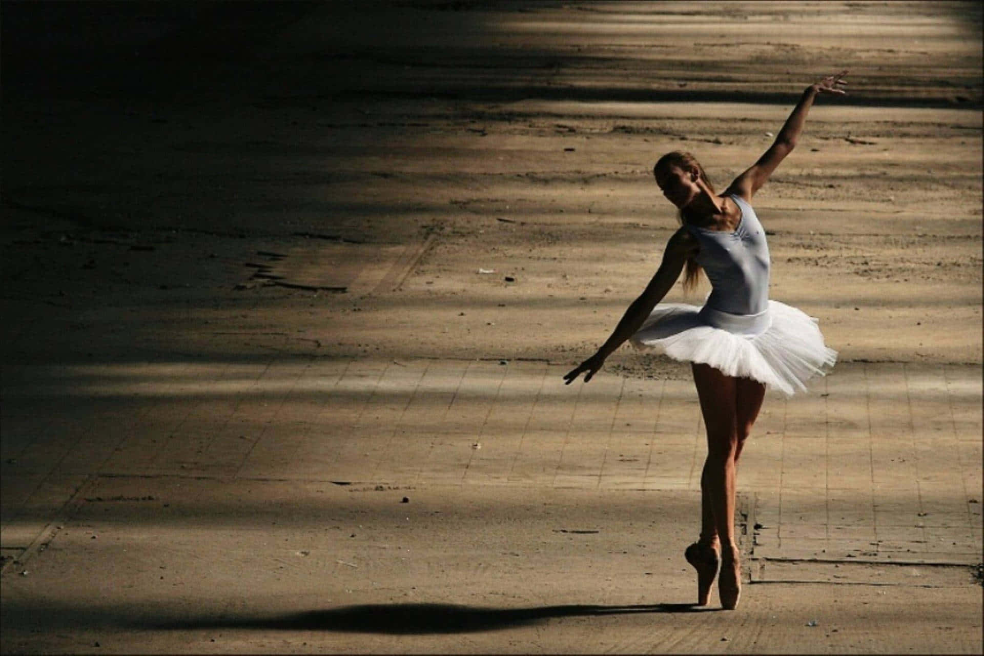 Ballet 桌布 壁紙 — 一位穿著白色芭蕾舞裙的芭蕾舞者，在陽光普照的混凝土廣場上優雅地踮起腳尖，雙臂伸展成優雅的阿拉貝斯克姿勢。