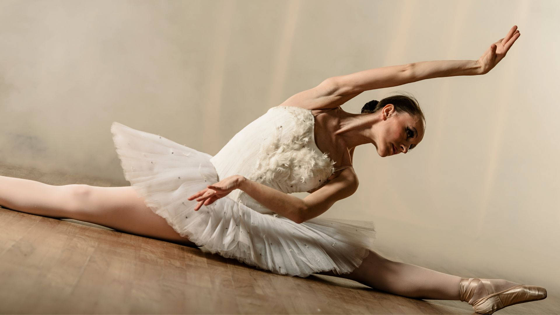 ballerina 桌布 壁紙 — 一位穿著白色芭蕾舞裙的芭蕾舞者在木地板上優雅地做出劈腿動作，背景是柔和的米色。