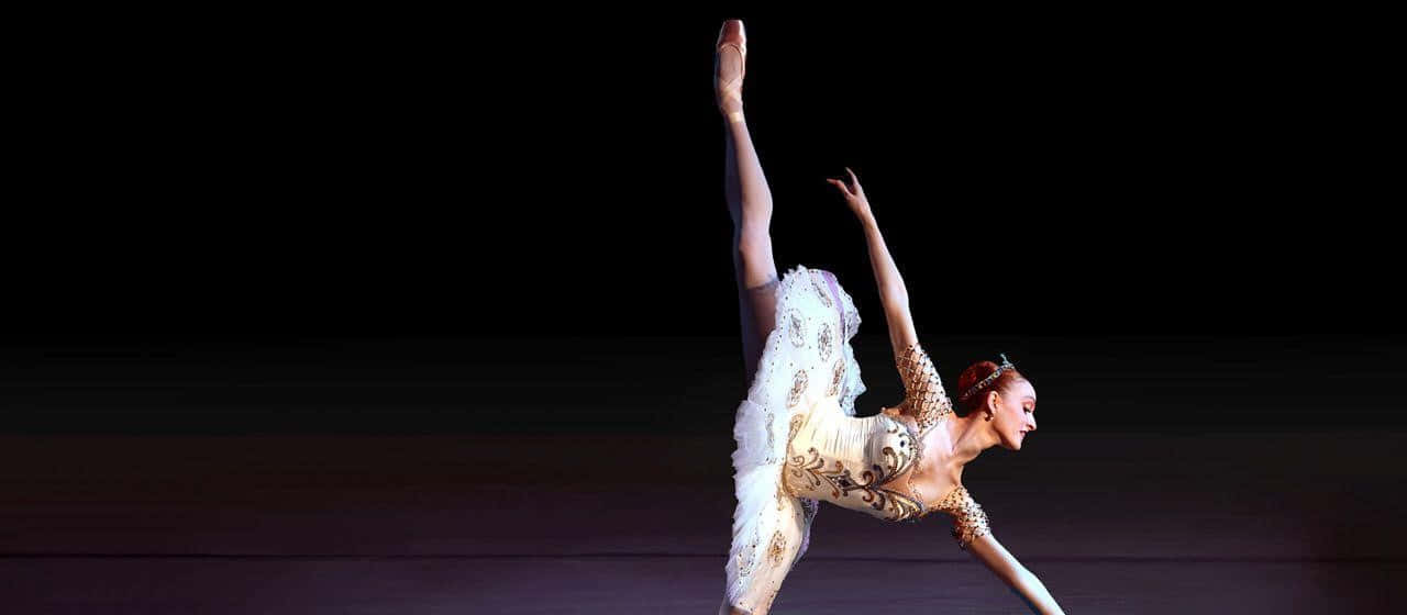 Ballet Dancer 桌布 壁紙 — 一位穿著白色芭蕾舞裙的芭蕾舞者在黑暗的舞台背景下表演優雅的阿拉貝斯克。