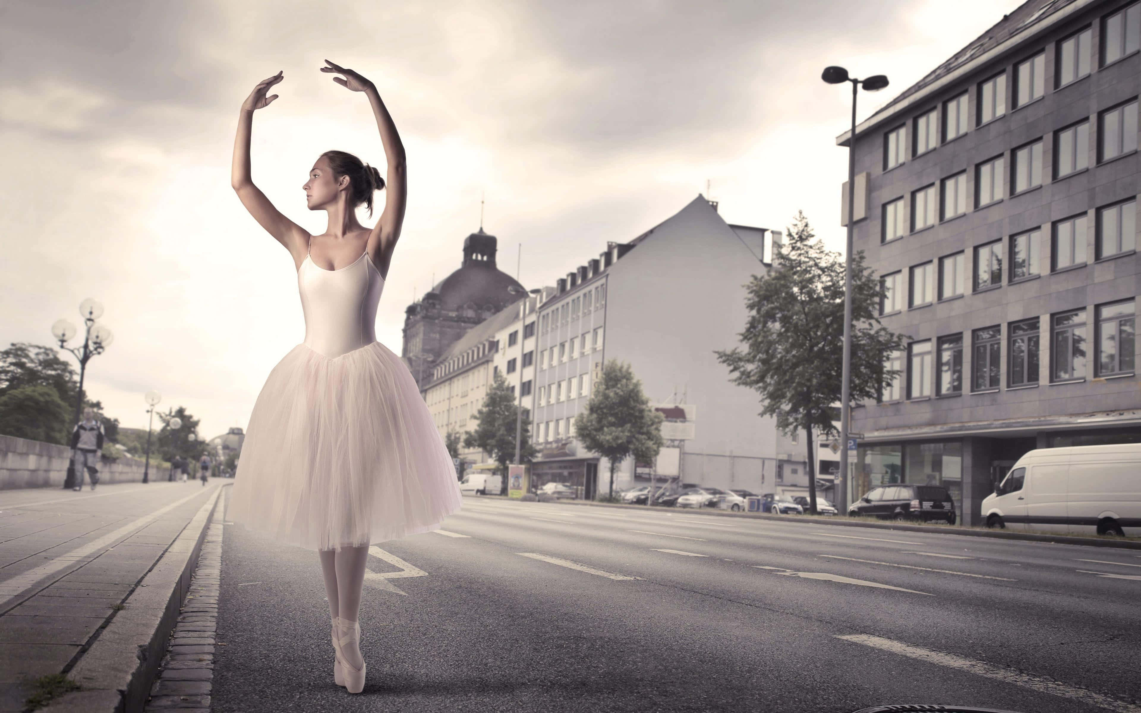 Ballet 桌布 壁紙 — 一位穿著白色芭蕾舞裙的芭蕾舞者在城市街道上優雅地踮起腳尖擺姿勢，背景有建築物和汽車。