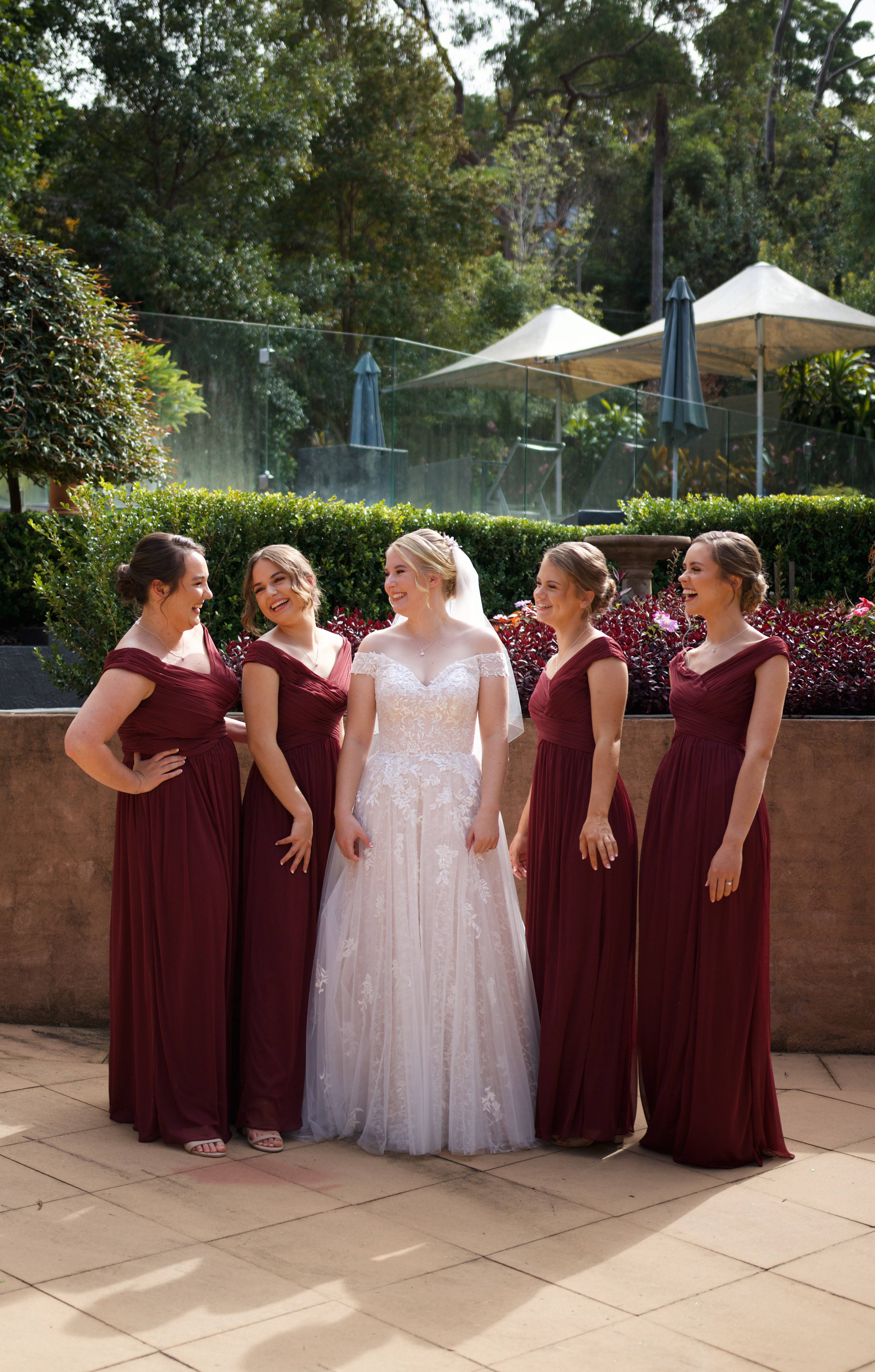 Bridesmaids 桌布 壁紙 — 一位穿著白色蕾絲婚紗的新娘與四位穿著相配酒紅色禮服的伴娘在戶外一起大笑。