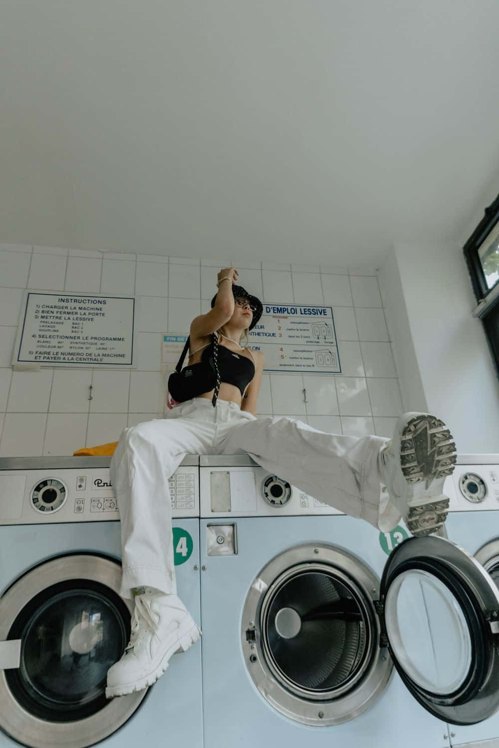 Laundry Room 桌布 壁紙 — 一位穿著白色褲子和靴子的時尚人士，懶洋洋地坐在開放式洗衣機上，身處鋪滿磁磚的洗衣店，牆上有法文指示。