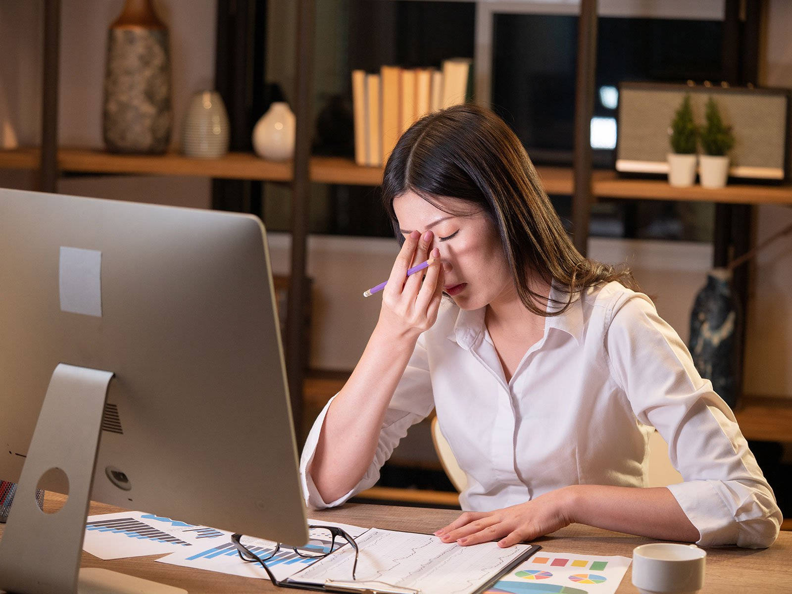 job 桌布 壁紙 — 一位穿著白色襯衫的疲憊女性，坐在現代辦公室裡堆滿圖表和電腦的凌亂辦公桌前，揉著眼睛。