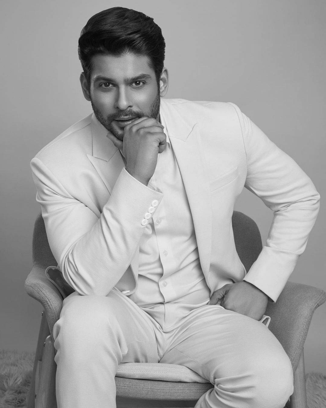 Sidharth Shukla 桌布 壁紙 — 一位穿著白色西裝、打扮俐落的男士自信地坐在椅子上，手托下巴，呈現極簡風格的攝影棚肖像。