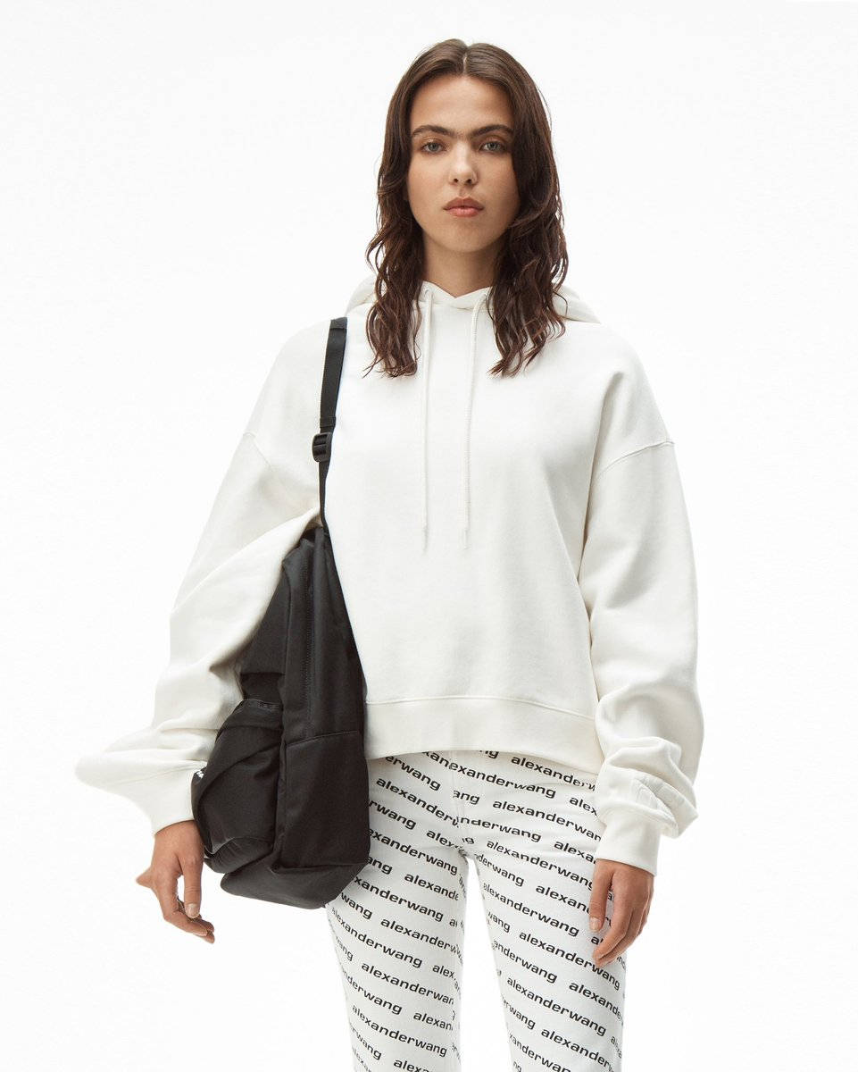 Alexander Wang 桌布 壁紙 — 一位穿著白色連帽衫和品牌緊身褲的女性，在純白色背景前背著一個黑色斜背包。