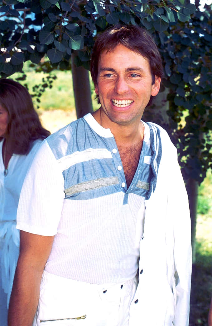 John Ritter 桌布 壁紙 — 一位穿著白藍條紋襯衫的開朗男子，在陽光明媚的日子裡，在綠樹成蔭下燦爛地微笑。