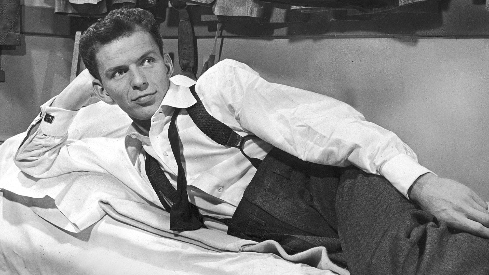 Sinatra 桌布 壁紙 — 一位穿著白襯衫和吊帶褲的時尚男士，在床上悠閒地躺著，表情若有所思。