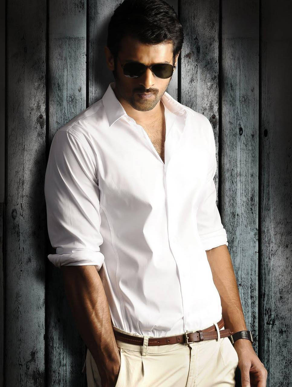 Prabhas Mirchi 桌布 壁紙 — 一位穿著白襯衫和太陽眼鏡的時尚男士，以自信的姿態靠在一面風化的木牆上。