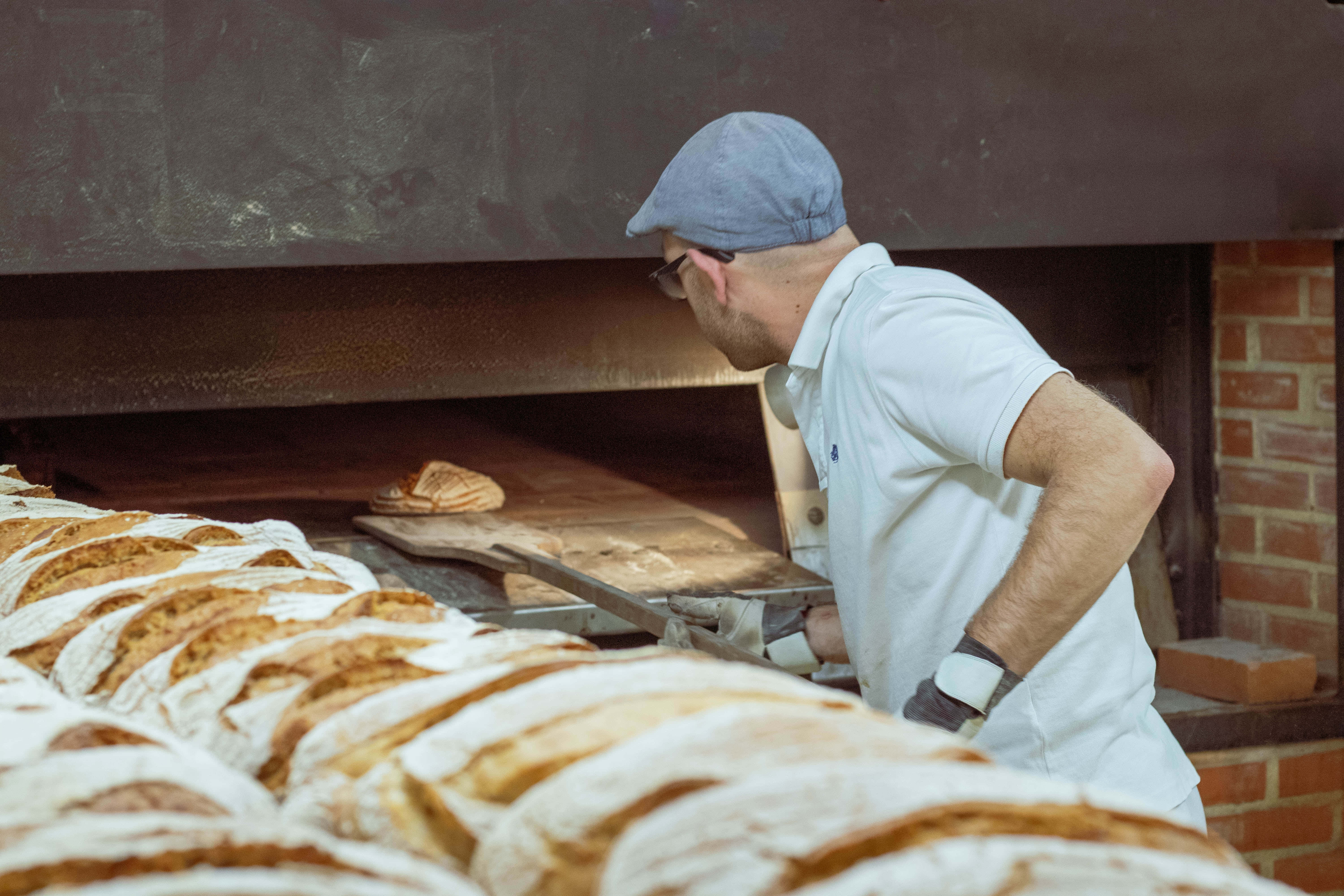 Bakery 桌布 壁紙 — 一位穿著白襯衫和藍色帽子的麵包師傅，小心翼翼地從鄉村風格麵包店的磚爐中取出金黃色的麵包。