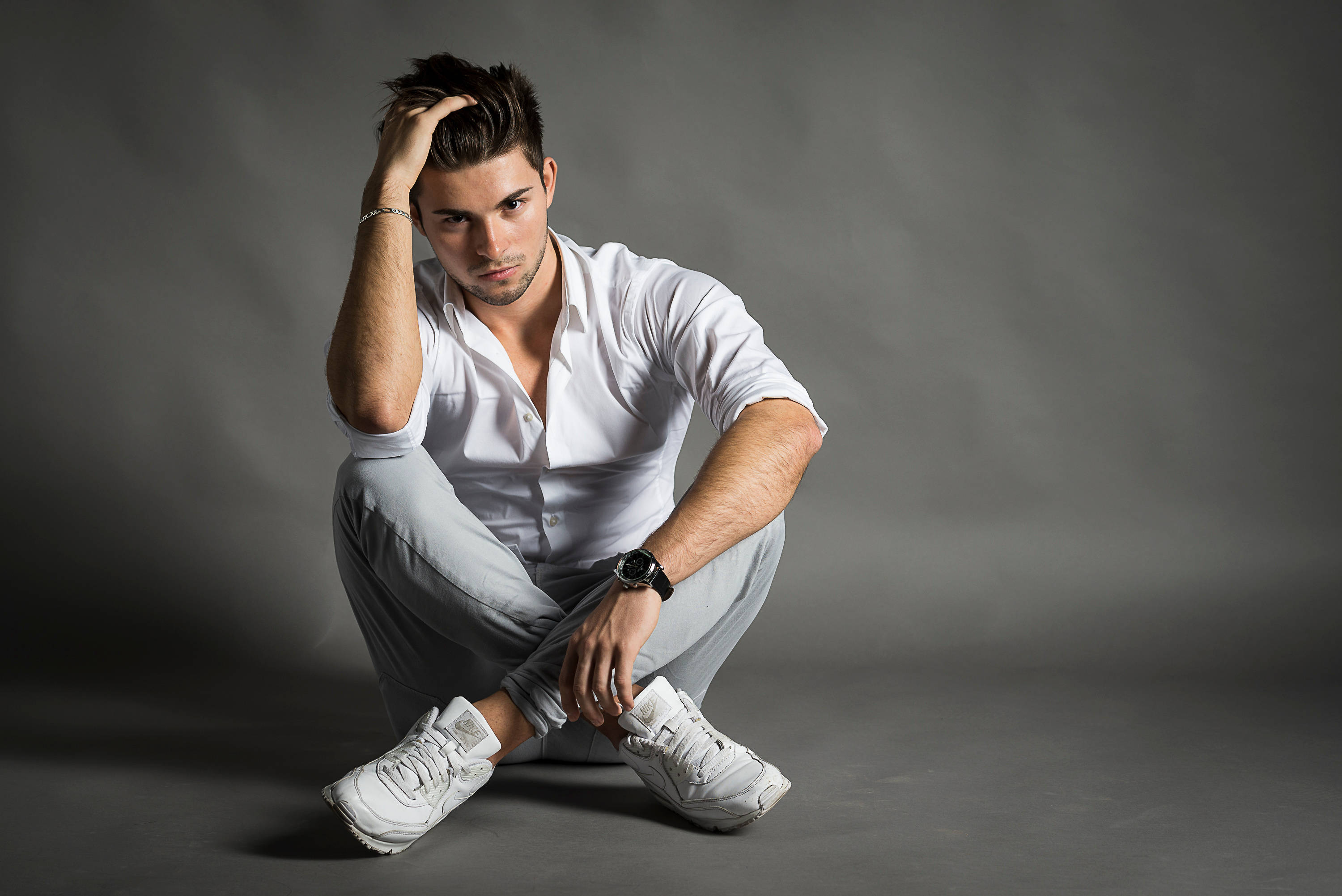 sexy man 桌布 壁紙 — 一位穿著白襯衫和運動鞋的時尚男士，在灰色攝影棚背景前蹲下，一隻手放在頭髮上。