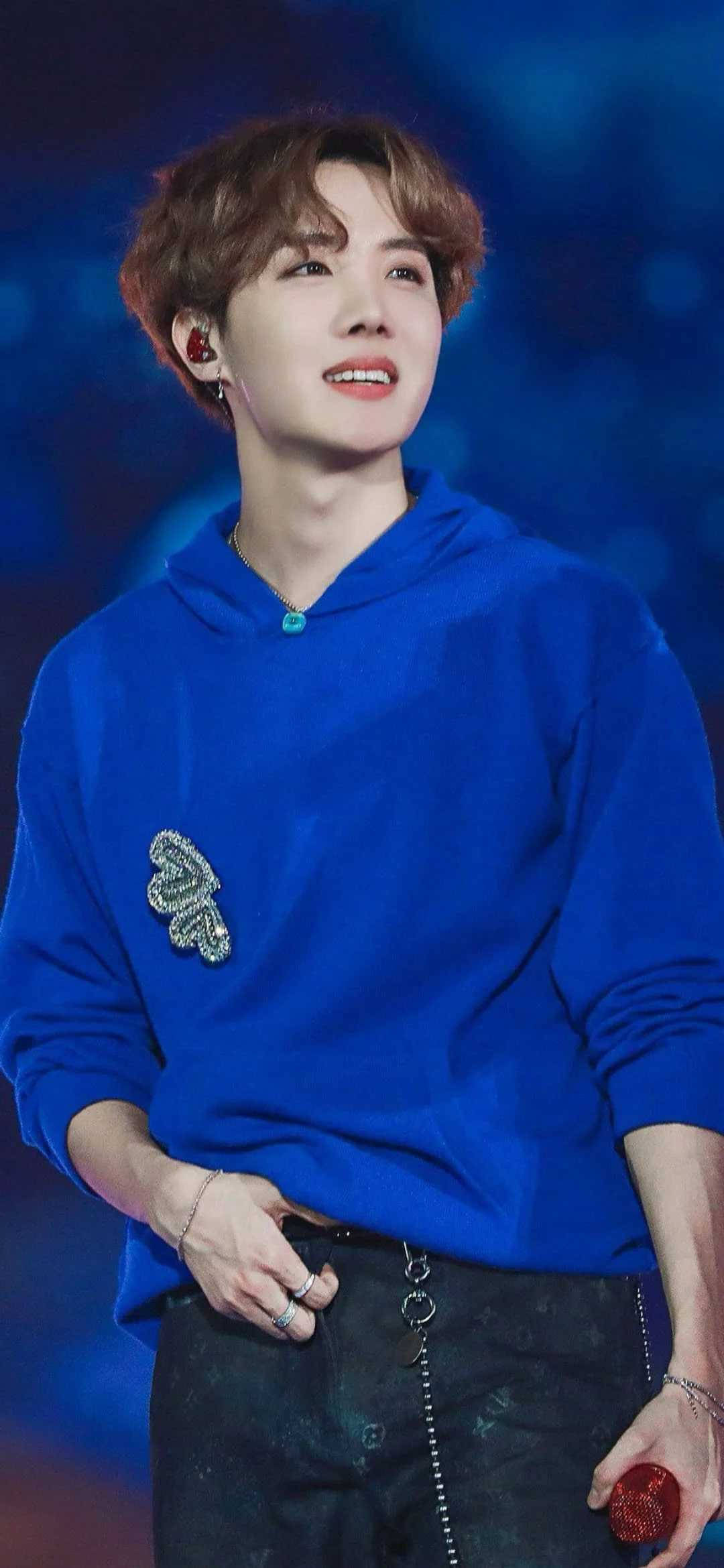 jhope 桌布 壁紙 — 一位穿著皇家藍色連帽衫的年輕男子在充滿活力的藍色舞台燈光下溫暖地微笑，同時手持紅色麥克風。