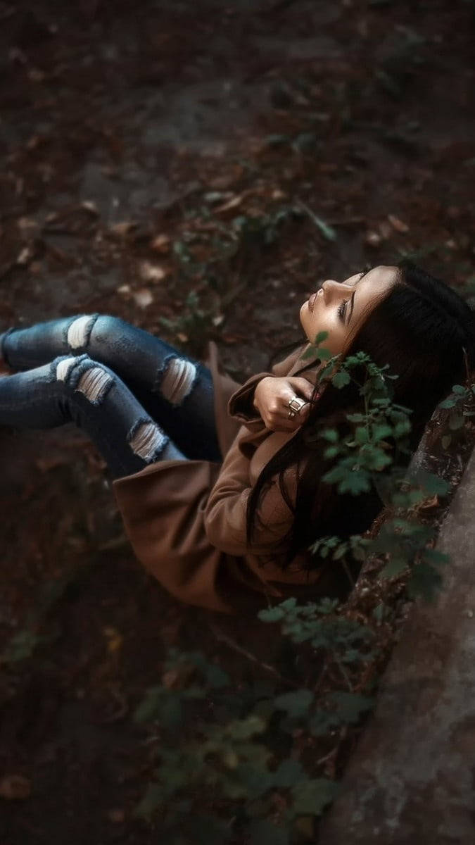 森林 桌布 壁紙 — 一位穿著破洞牛仔褲和棕色外套的年輕女性躺在森林地面上，周圍環繞著落葉和綠色藤蔓。