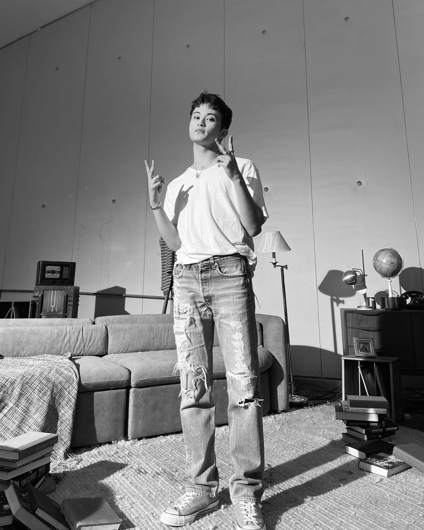 Nct 桌布 壁紙 — 一位穿著破洞牛仔褲和白色T恤的年輕男子，在陽光充足的簡約房間裡自信地站著，比出和平手勢。
