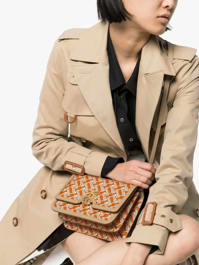 Burberry 桌布 壁紙 — 一位穿著米色風衣的女性，在純白色背景前拿著一個橙色圖案的手提包。