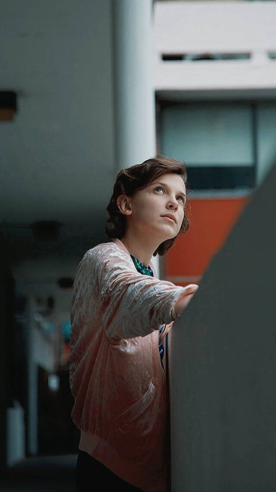 Millie 桌布 壁紙 — 一位穿著粉紅色外套的年輕人靠著牆壁向上看，身處一個色調柔和的現代室內空間。