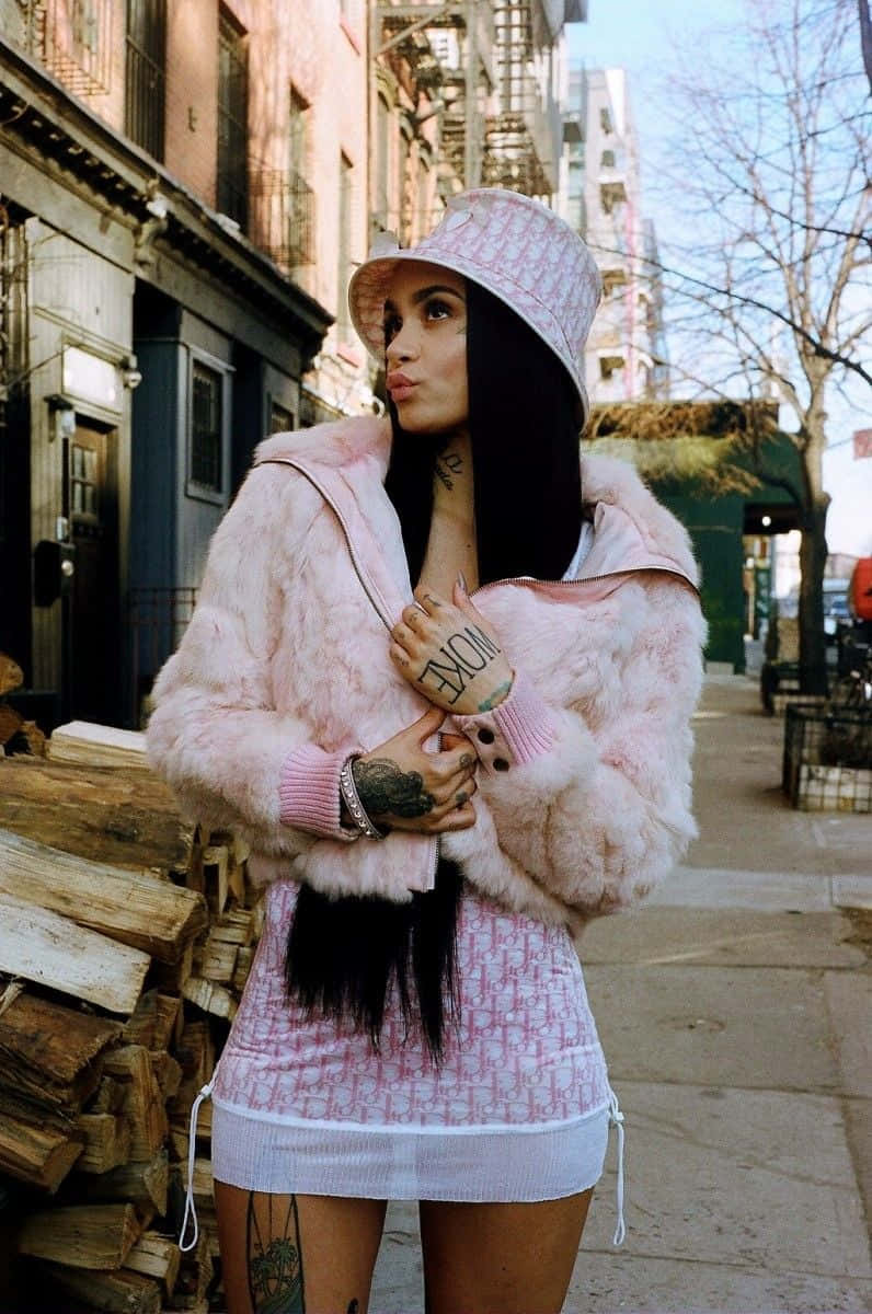 kehlani 桌布 壁紙 — 一位穿著粉紅色毛皮大衣和配套漁夫帽的女性自信地站在城市人行道上，旁邊是堆疊的木柴。