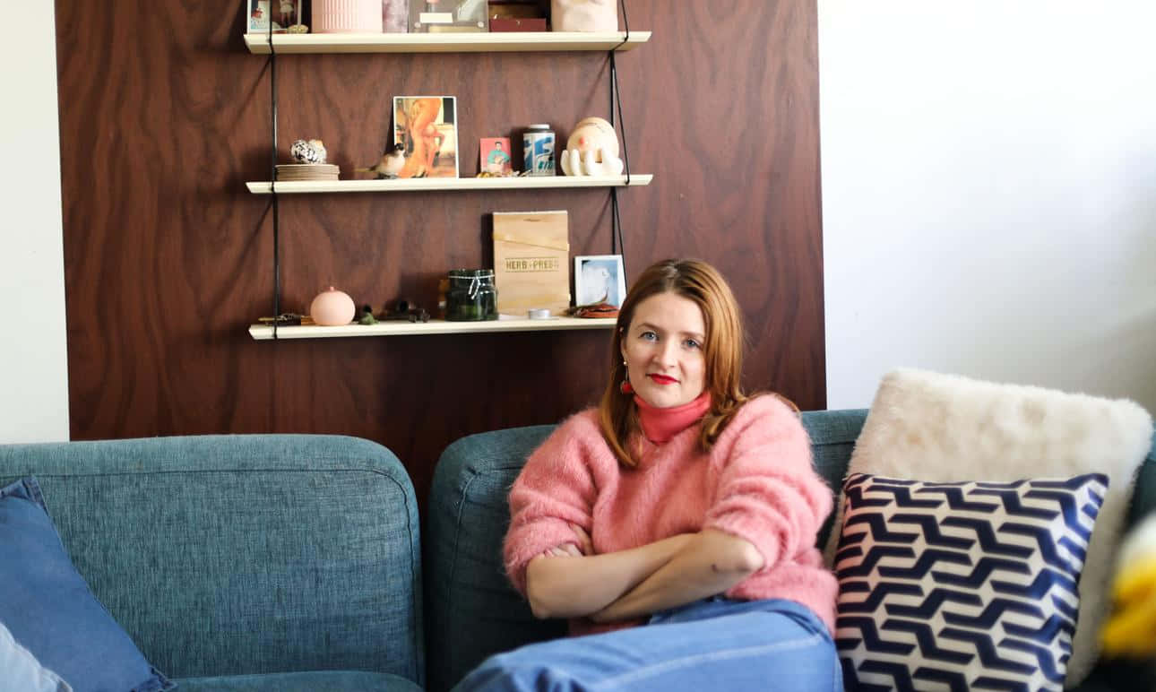 Woman 桌布 壁紙 — 一位穿著粉紅色毛衣的女性自信地坐在藍色沙發上，沙發上有圖案靠墊，背景是木質層架牆。