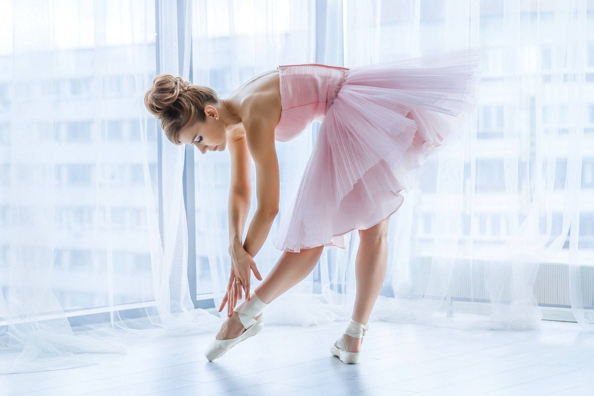 Ballet 桌布 壁紙 — 一位穿著粉紅色芭蕾舞裙的芭蕾舞者在陽光充足的舞蹈室中優雅地伸展，室內有薄紗窗簾和城市景觀。