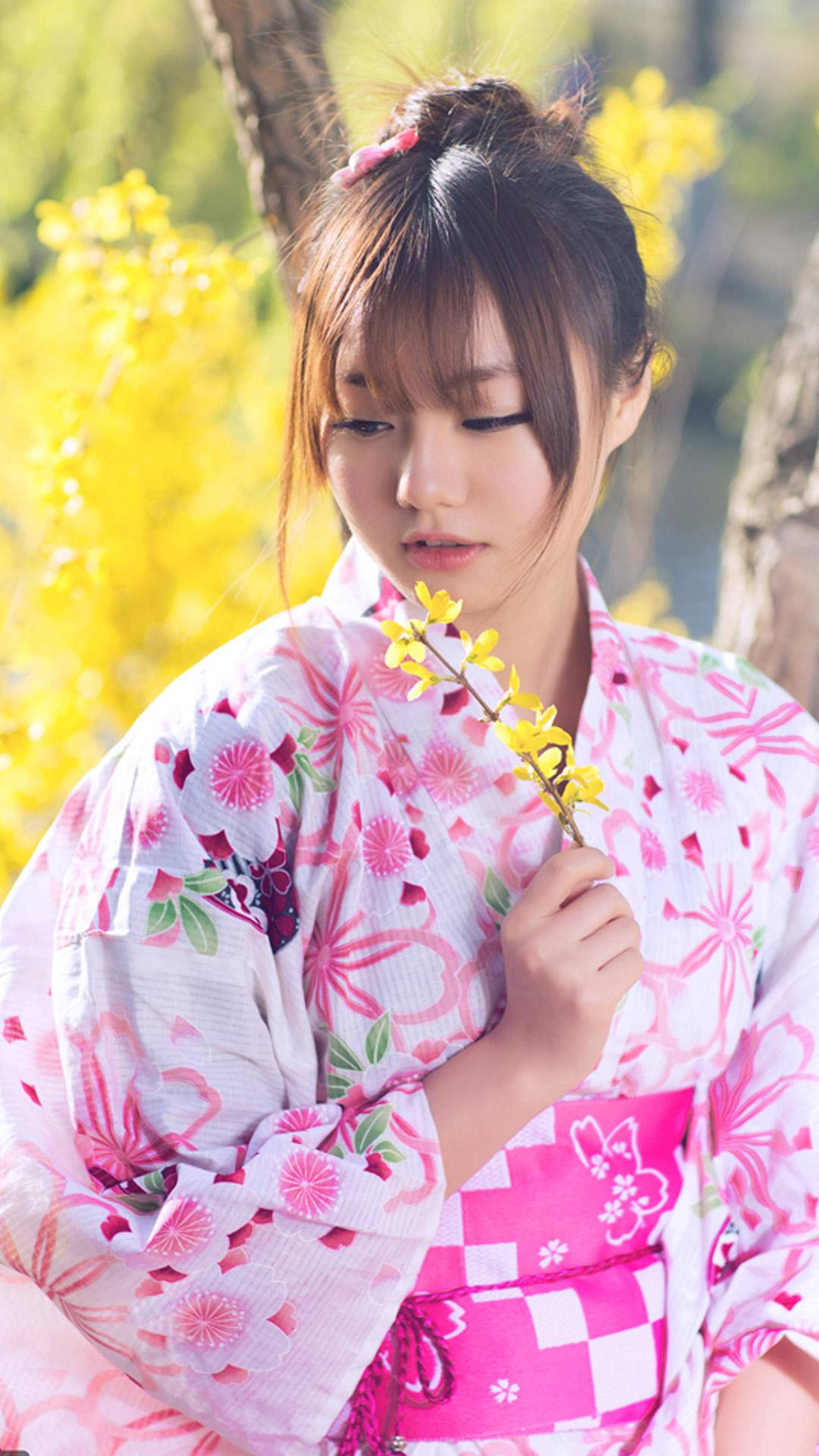 日本 桌布 壁紙 — 一位穿著粉紅色花卉和服的年輕女子，在陽光明媚的春天背景下手持黃色花枝。