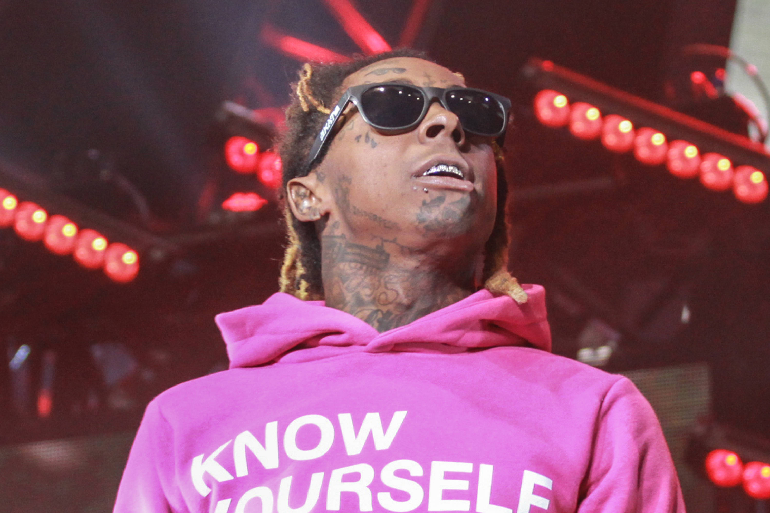 Lil Wayne 桌布 壁紙 — 一位穿著粉紅色連帽衫和太陽眼鏡的饒舌歌手在紅色舞台燈光下表演，身上有可見的刺青。