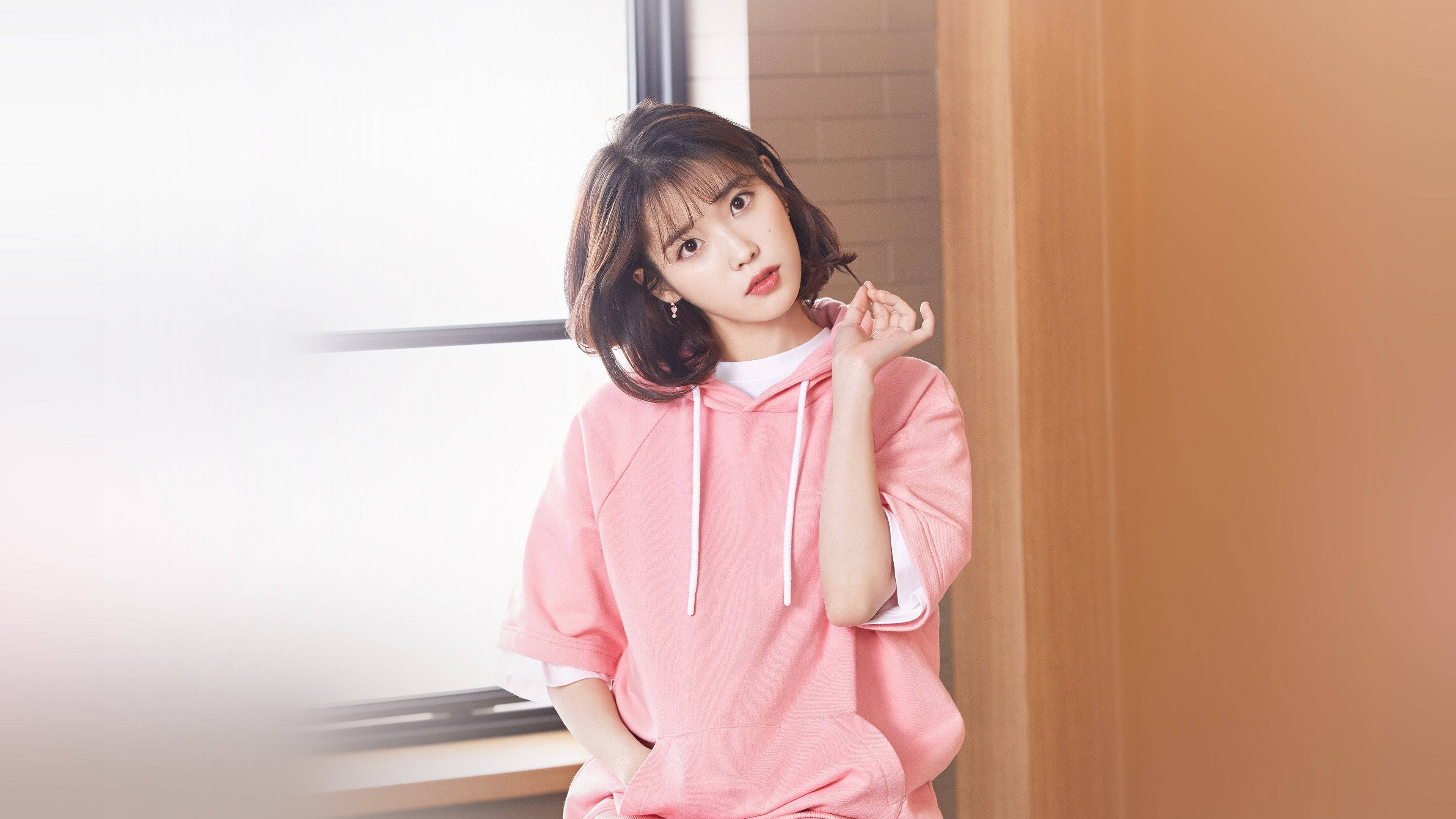 Kpop Aesthetic 桌布 壁紙 — 一位穿著粉紅色連帽衫的年輕女子靠著木門框，身後窗戶透進柔和自然光。