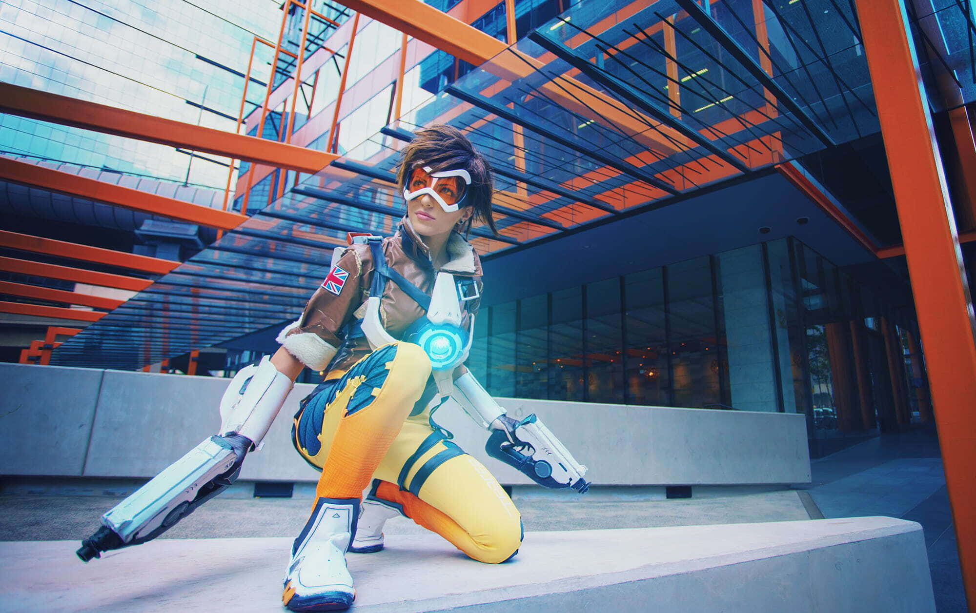 Gaming Cosplay 桌布 壁紙 — 一位穿著精細《鬥陣特攻》閃光服裝的角色扮演者，在現代玻璃與橙色鋼結構建築外牆前動態蹲下。