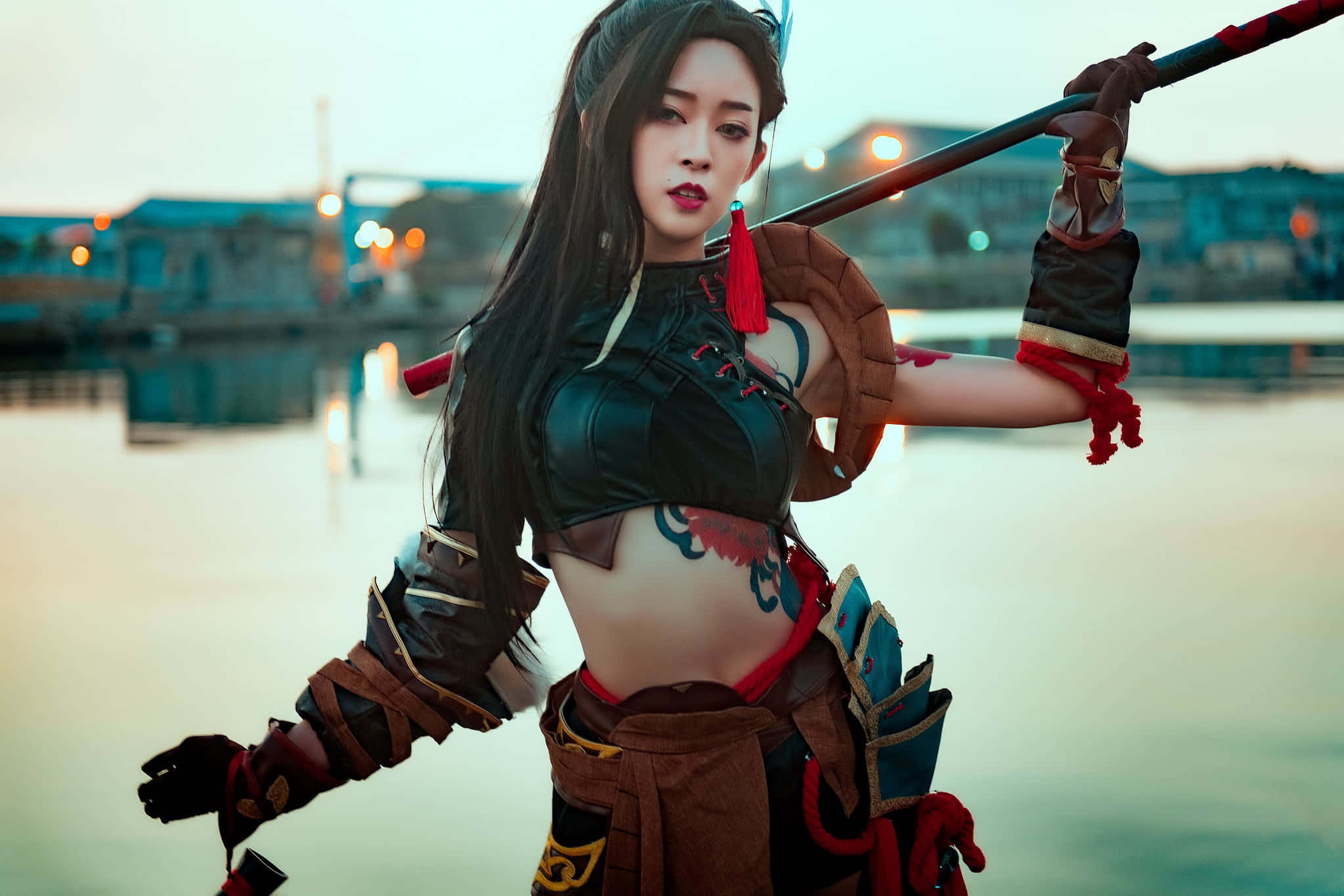 Gaming Cosplay 桌布 壁紙 — 一位穿著精細戰士服裝的角色扮演者在黃昏時分於水邊手持法杖擺姿勢，背後是模糊的城市燈光。