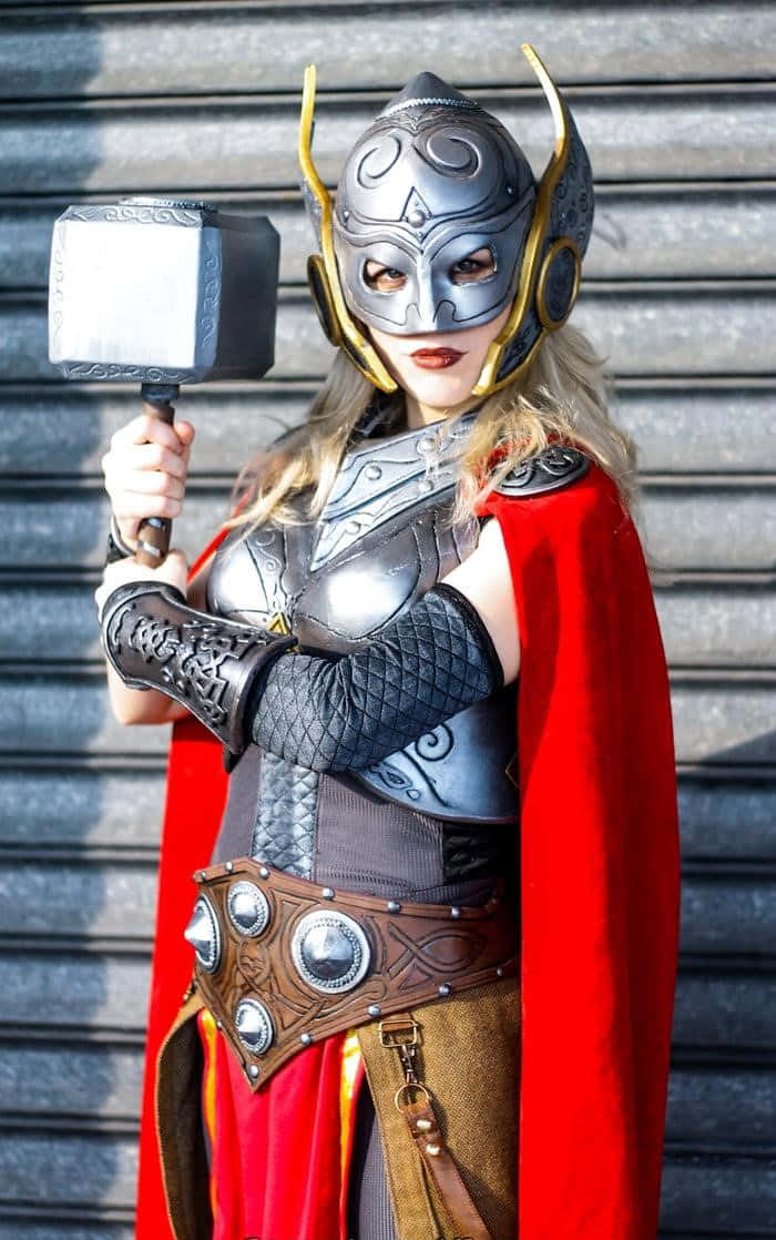 Jane Foster 桌布 壁紙 — 一位穿著精細雷神服裝的角色扮演者在明亮的日光下，在波紋金屬背景前舉著錘子。