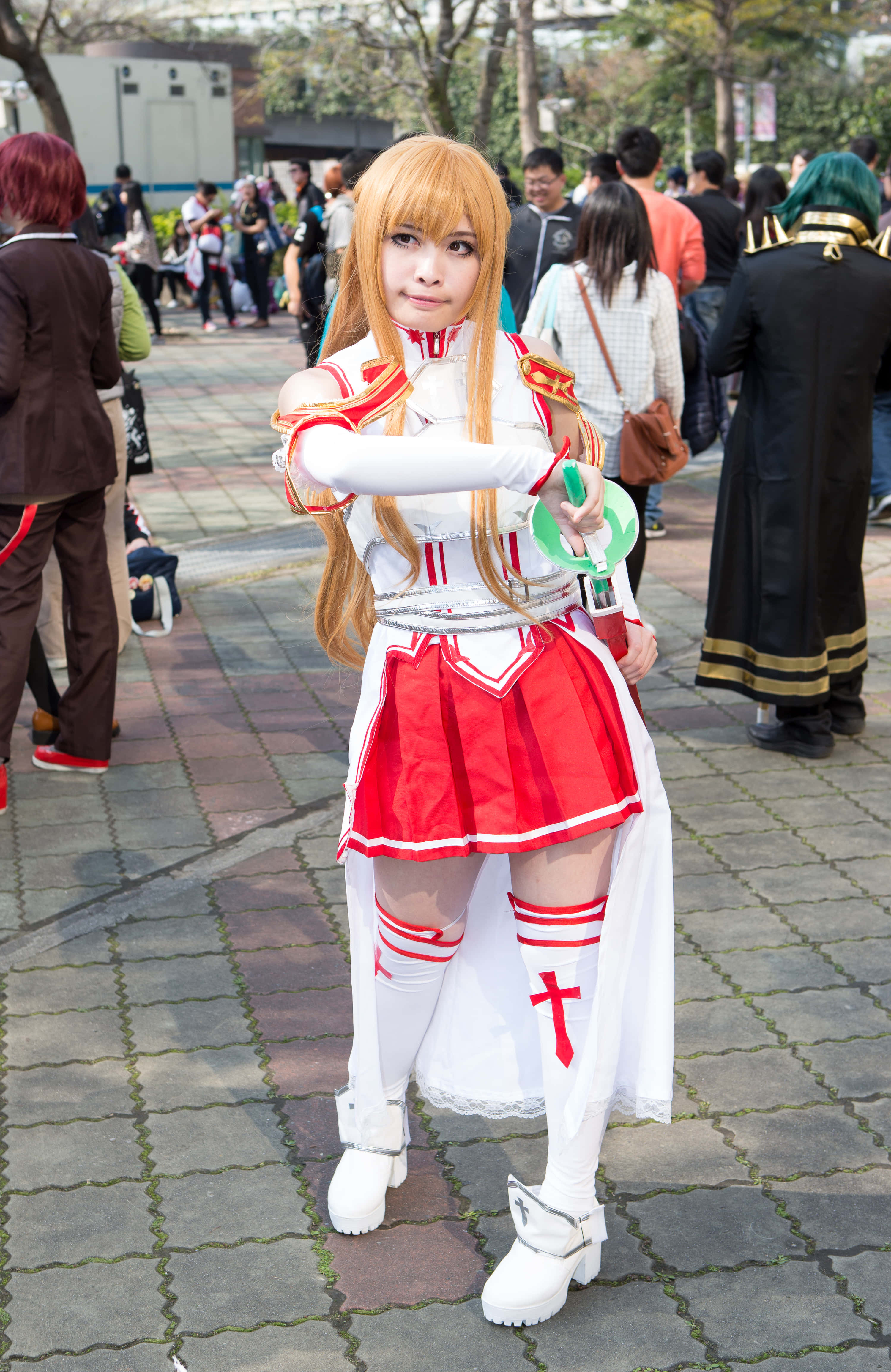Sword Art Online Cosplay 桌布 壁紙 — 一位穿著紅白奇幻服裝的角色扮演者在鋪砌的戶外走道上自信地擺姿勢，周圍有其他參與者。