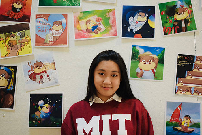 學校 桌布 壁紙 — 一位穿著紅色MIT運動衫的年輕女子對著鏡頭微笑，站在一面佈滿色彩繽紛卡通藝術的牆前，牆上有一個重複出現的熊角色。