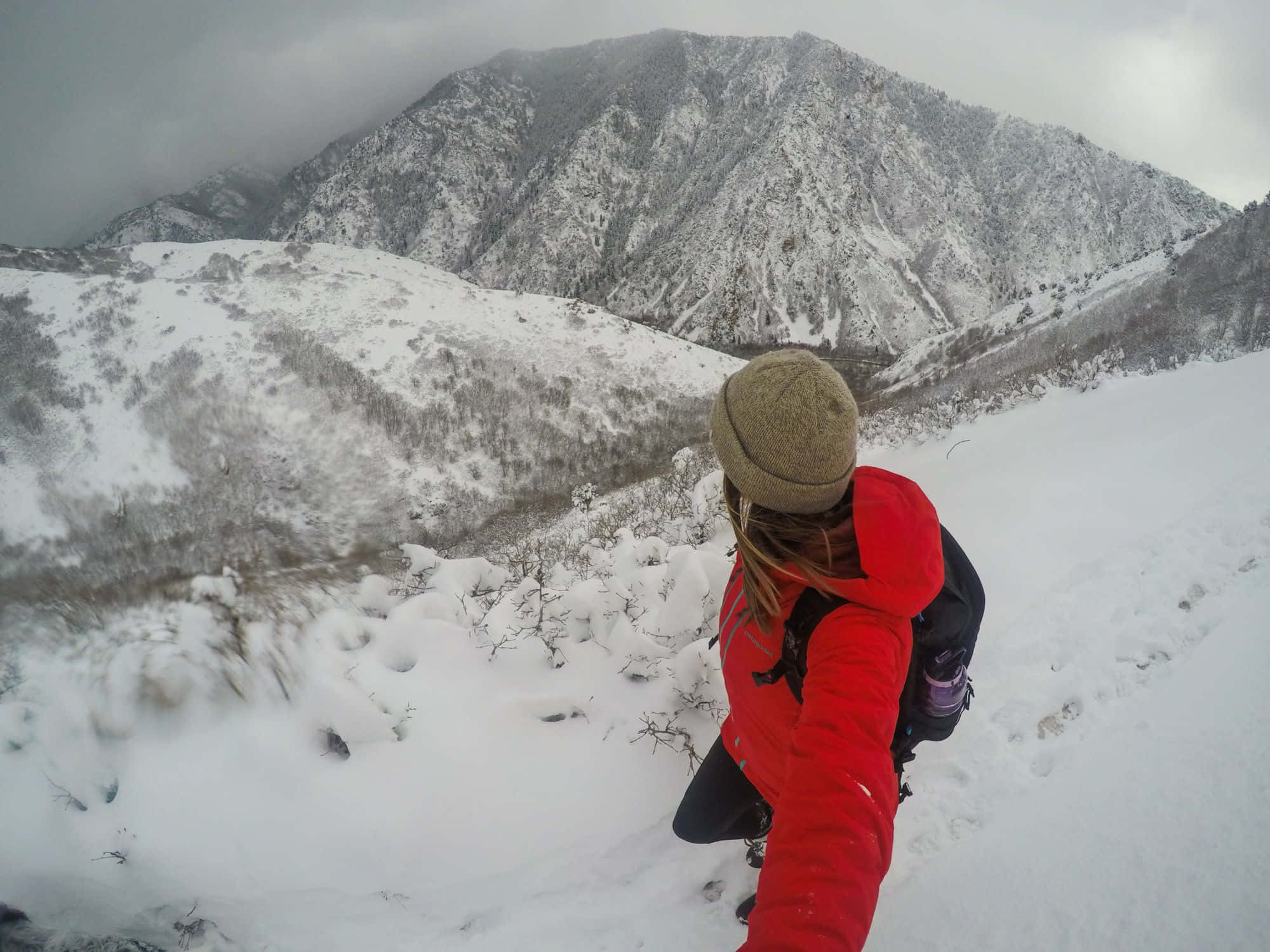 Winter Hiking 桌布 壁紙 — 一位穿著紅色外套和毛帽的登山者在多雲的天空下穿越白雪覆蓋的山谷時自拍。