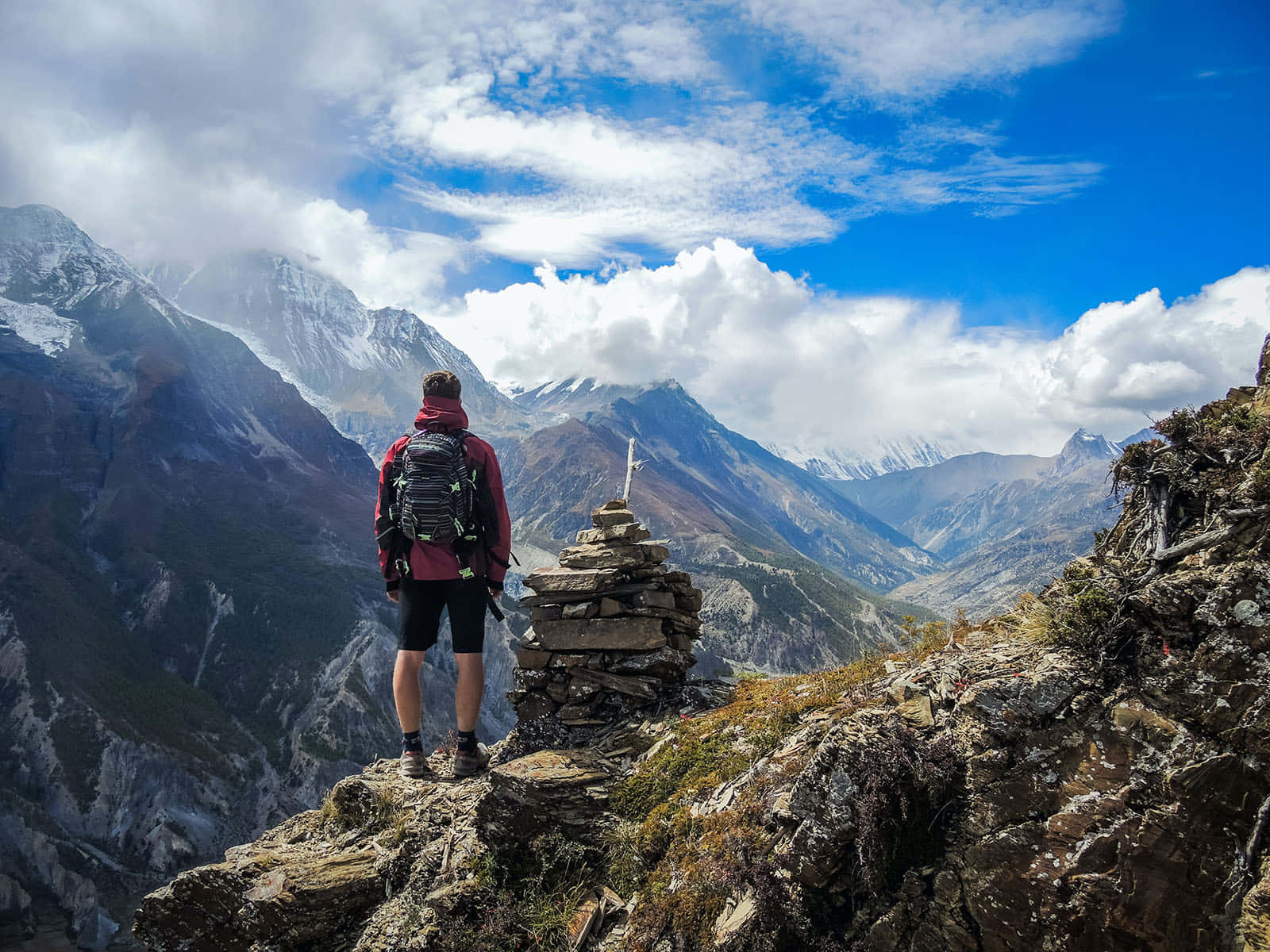 Traveling 桌布 壁紙 — 一位穿著紅色夾克的徒步旅行者站在岩石懸崖上，俯瞰著白雪覆蓋的山脈，天空湛藍，白雲朵朵。